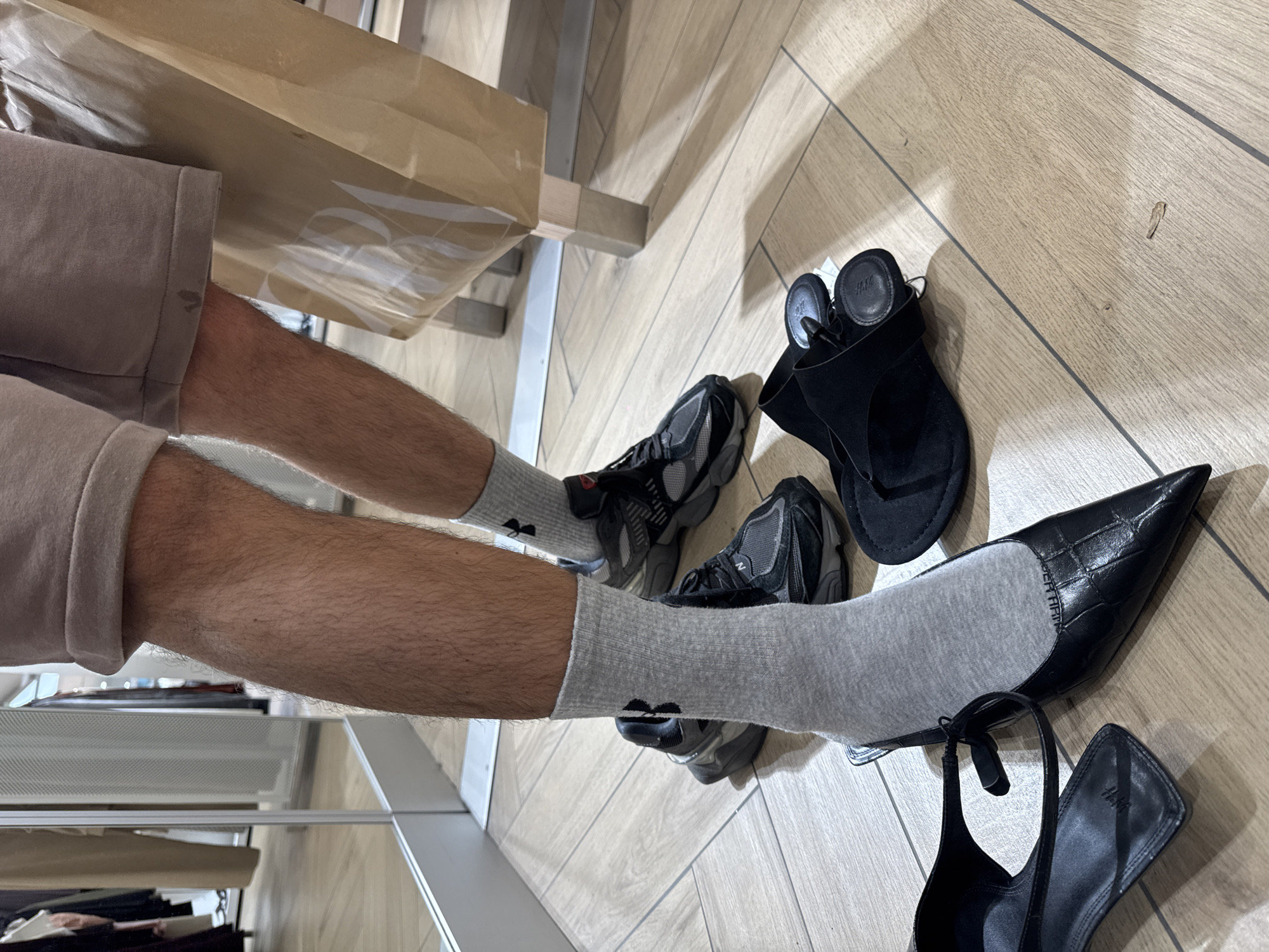 The image depicts a close-up view of a person's lower legs and feet within a retail store or fitting room in Plovdiv, Bulgaria. The individual, possessing visibly hairy legs, is wearing cuffed light brown shorts and grey ankle socks, each featuring a black Under Armour logo. The right foot (viewer's left) is currently wearing a black pointed-toe heel with a textured finish and an ankle strap, worn over the sock. On the light-colored wooden tiled floor, a pair of black and grey New Balance 9060 sneakers are positioned behind the feet. Adjacent to the worn heel, another identical black pointed-toe heel, marked with an H&M logo on its inner sole, lies on the floor. Additionally, a pair of black open-toe sandals, one displaying an H&M logo on its sole, are also present on the floor. To the right, a large brown paper shopping bag with a partially visible ZARA logo is standing. The background features a large mirror reflecting racks of clothing and garments, characteristic of a retail environment.