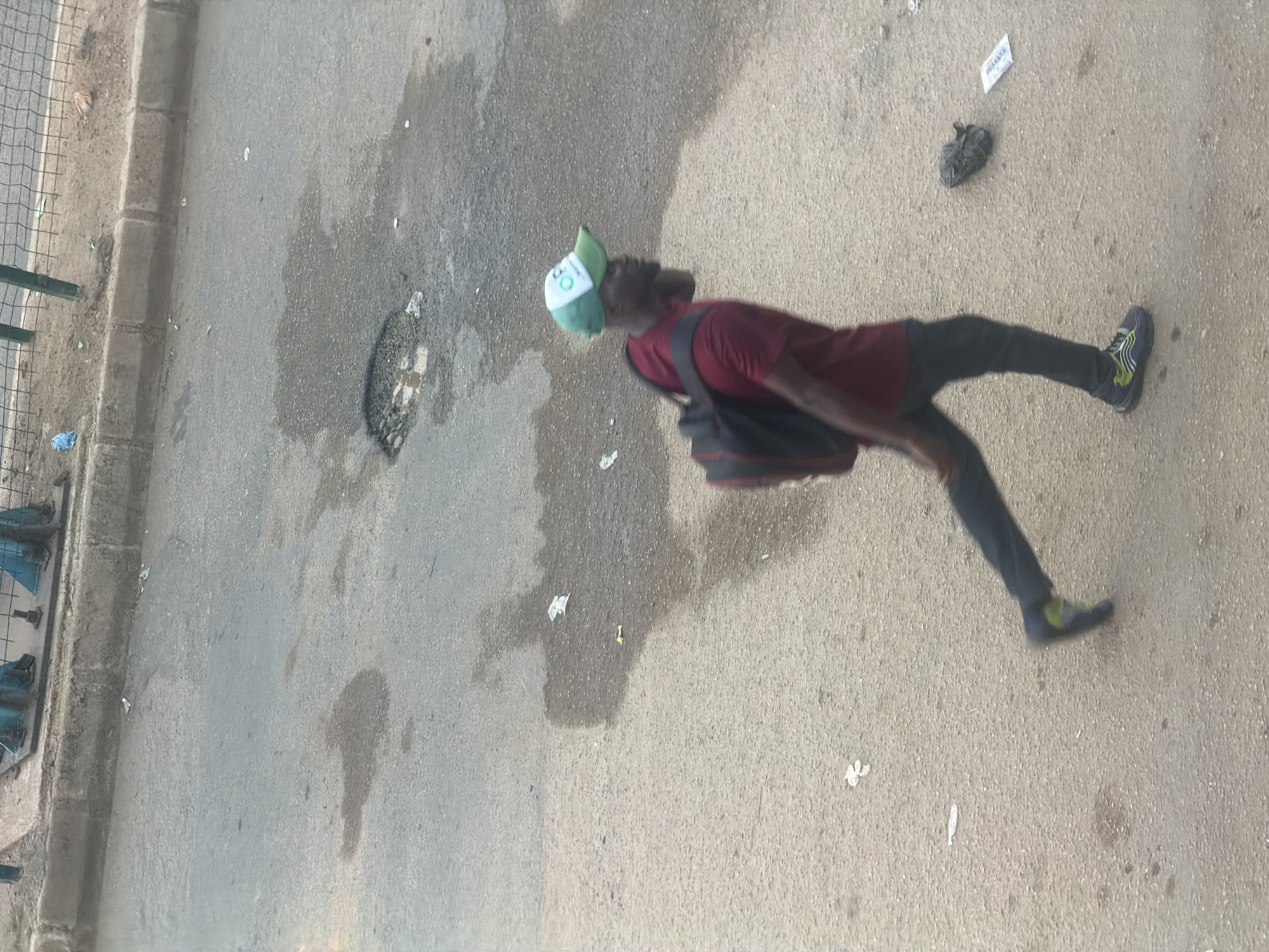 The image shows a person walking on a paved road in Bukuru, Nigeria. The person is wearing a maroon t-shirt, dark pants, a green and white baseball cap, and sneakers. They are also carrying a dark bag slung over their shoulder. The road surface appears to be asphalt and is wet in some areas, suggesting recent rain or a spill. There is some litter on the ground, including a plastic bag and what looks like a crumpled piece of paper. The scene is outdoors, and the lighting suggests it is daytime. The person's body language does not convey any strong emotions. In the background, there is a low barrier, possibly part of a fence or railing, and some urban debris. There are no clear indicators of the time of day or specific weather conditions beyond the wet road.