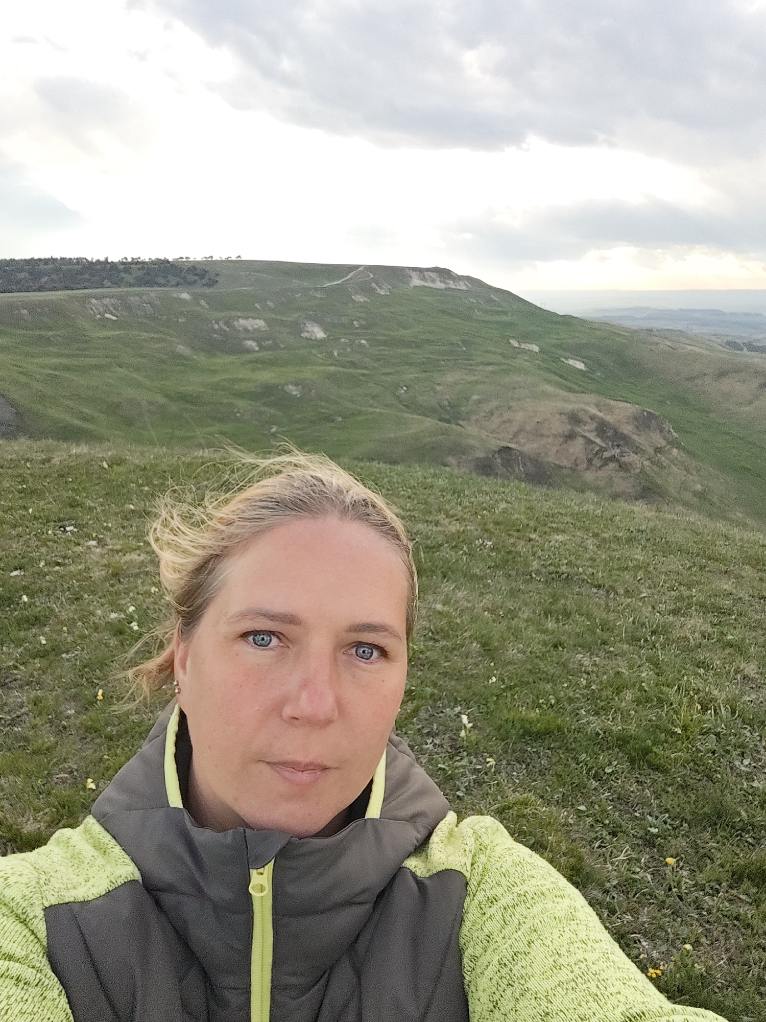 A woman with blonde hair and blue eyes is the primary subject of the image. She is wearing a gray and neon green jacket with a visible zipper. The background consists of a green, hilly landscape beneath a cloudy sky. In the distance, there is a distinct plateau-like hill formation with what appears to be a path leading to its summit. The location is Kichmalka, Russia.