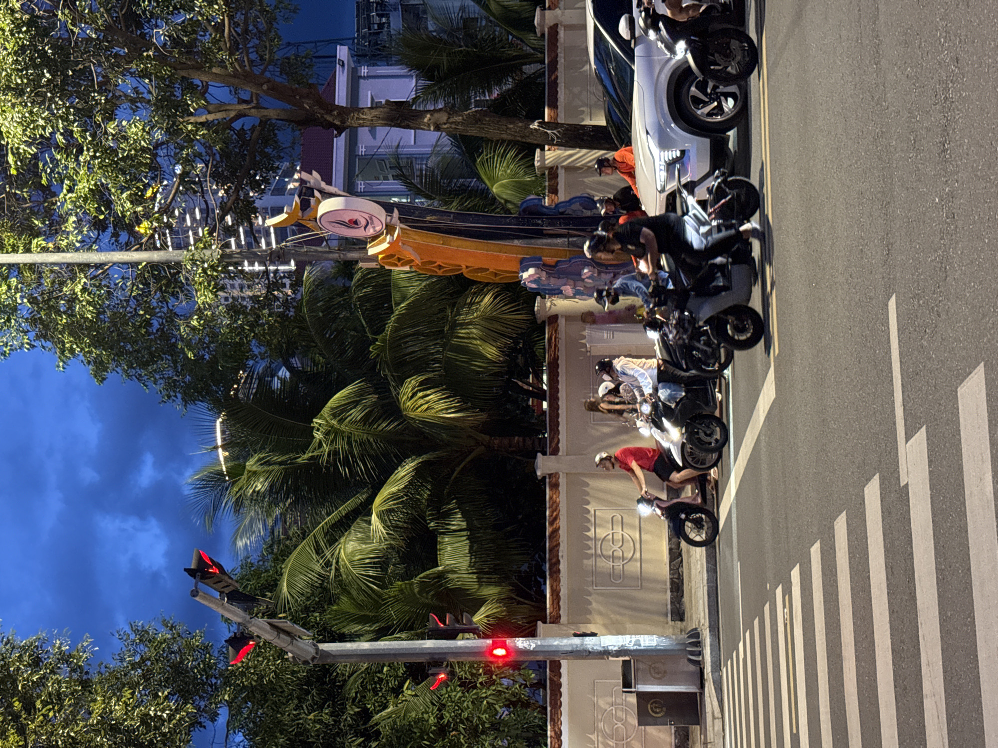 The image depicts a street scene in Nha Trang, Vietnam, at dusk or early night. A traffic light with a red signal is visible, indicating that vehicles should stop. Several people are on motorbikes, some waiting at the intersection, while others are in motion. A white SUV is also present. The background features palm trees and buildings, suggesting an urban environment. The street is marked with a crosswalk. The sky is a deep blue, indicating the transition from day to night.