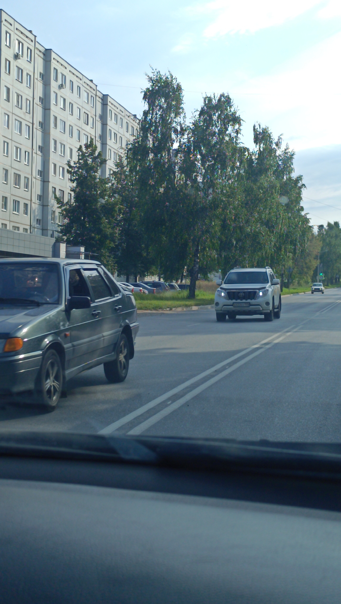 The image depicts a street scene in Chaikovsky, Russia. In the foreground, a dark green sedan is driving on a paved road with double white lane markings. The sedan's windows are tinted, and the interior is not clearly visible. Behind the sedan, a white SUV is also traveling on the road. Further in the background, a multi-story apartment building with numerous windows lines the left side of the street. Trees with dense green foliage are visible between the building and the road, and along the right side of the road. The sky is partly cloudy. The overall impression is a typical urban or suburban street on a bright day.