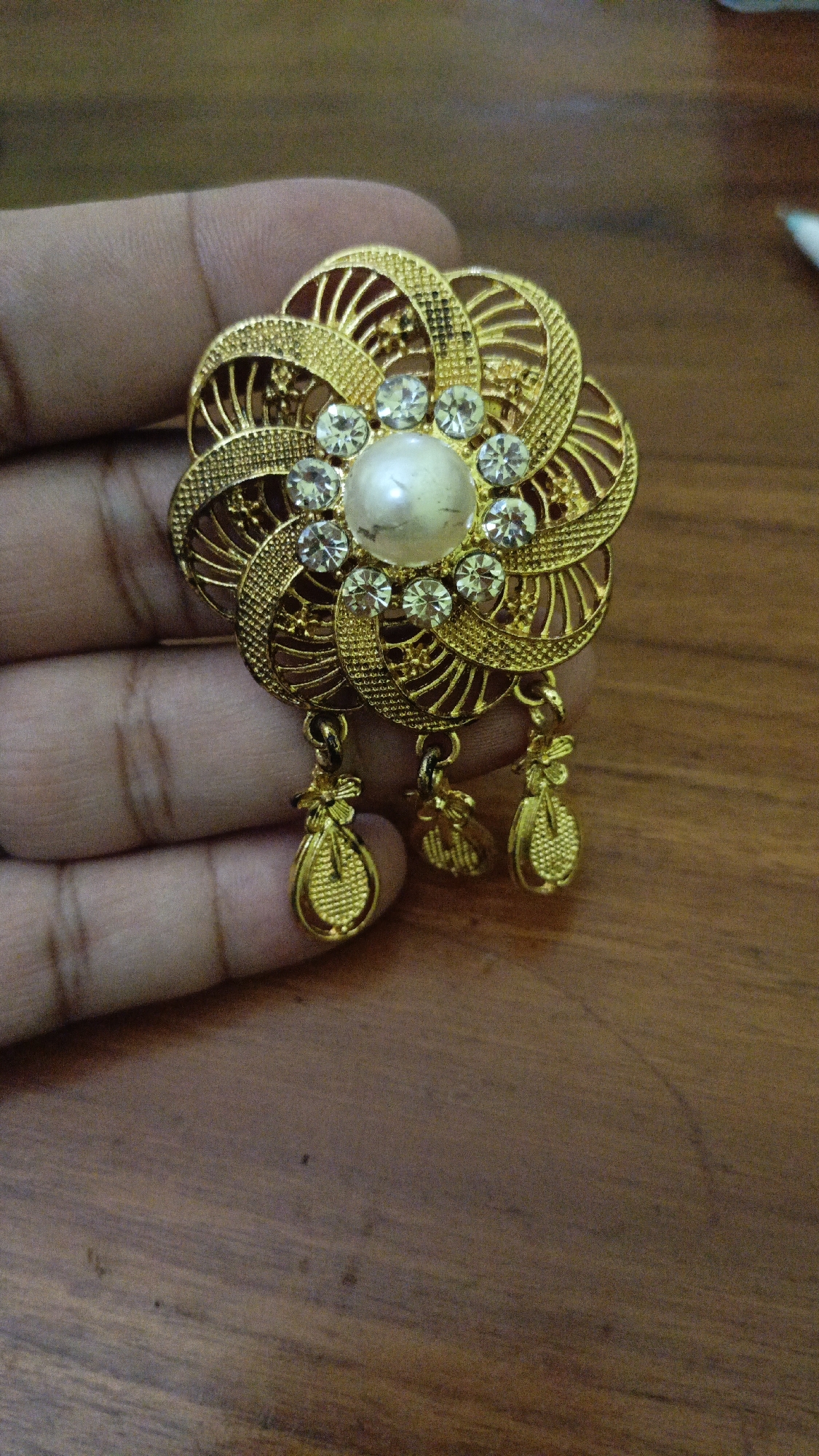 A hand holds a gold-colored ornate brooch featuring a central pearl surrounded by clear stones, with three pendant charms hanging from the bottom. The brooch is intricate, with a floral design and filigree work. The background is a wooden surface. The location is described as Trihanggo, Indonesia.