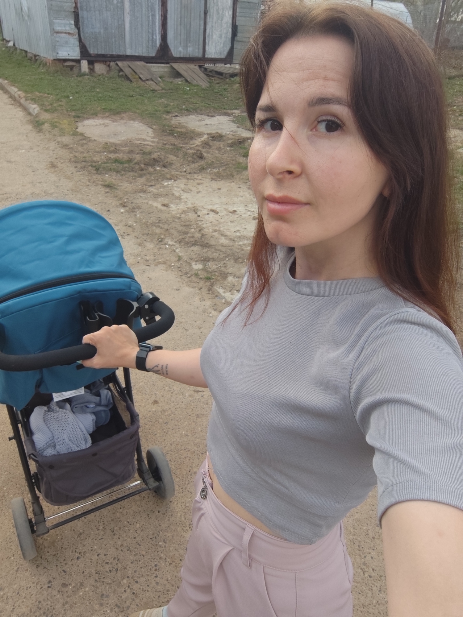 A woman is standing outdoors next to a baby stroller, taking a selfie. She has long brown hair and is wearing a grey crop top and pink pants. The stroller is blue and black, with items inside the storage compartment. The background consists of a dirt road and a dilapidated metal building. The location is Фоки, Russia.