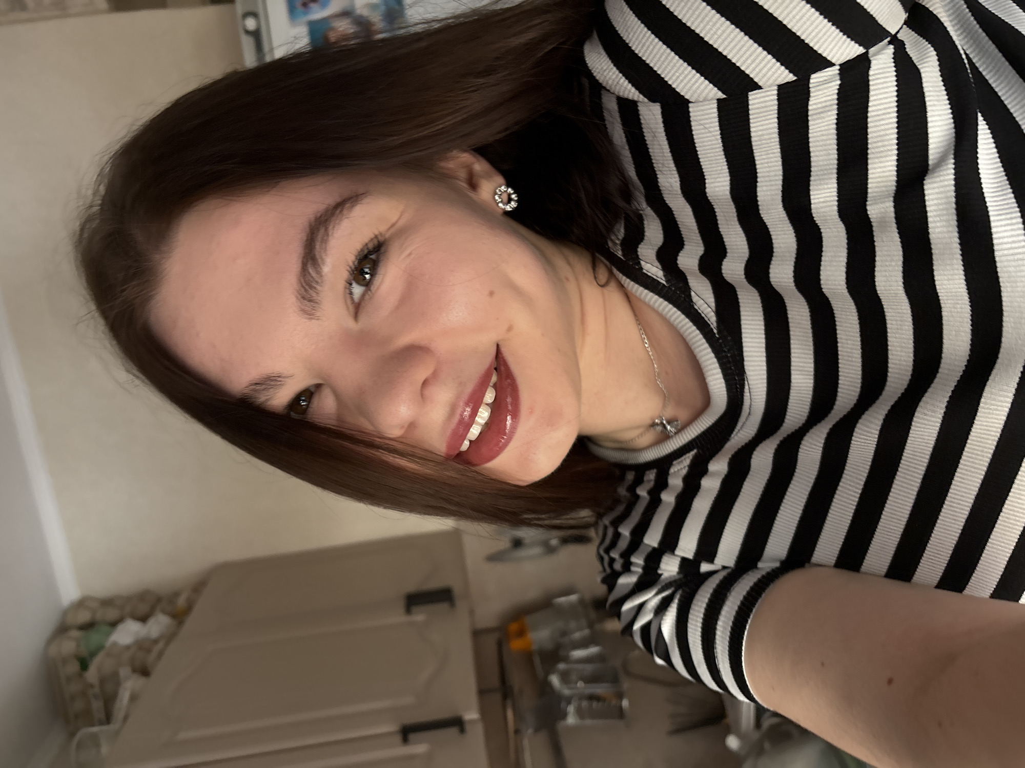 A young woman with dark brown hair, fair skin, and brown eyes is smiling slightly at the camera. She is wearing a black and white striped top and a silver necklace. She has earrings. The background shows a kitchen interior with light-colored cabinets and various items on shelves, indicating a domestic setting. The location is likely in Perm, Russia, but there is no specific confirmation within the image itself, only if that is where the photo was taken.