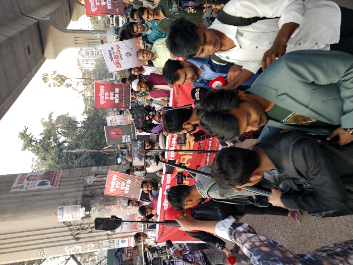 This image depicts a public gathering or demonstration on an urban street in Dhaka, Bangladesh, situated beneath a concrete elevated road or flyover structure. Approximately 20-30 individuals are visible in the foreground and midground, predominantly young to middle-aged males, dressed in casual to semi-formal attire.
In the midground, several individuals hold rectangular red placards with white Bengali text, notably displaying "ফুলবাড়ীয়ার" (Fulbariyar), "মুক্তি চায়" (wants freedom), and slogans such as "শহীদদের রক্ত বৃথা যাবে না" (the blood of martyrs will not go in vain). A long red banner with similar white Bengali text, including "স্মরণসভা" (commemoration meeting), is stretched across the front of the gathering. A central figure in a white cap and maroon waistcoat is positioned behind this banner, seemingly addressing the crowd or media.
Numerous individuals are actively documenting the event, utilizing smartphones mounted on tripods or selfie sticks. Additionally, several professional microphones with media logos (e.g., "MIRROR") are visible, held by journalists. The concrete pillar supporting the elevated road is covered with various posters and Bengali graffiti, including a poster featuring a person's image. The background reveals partial views of vehicles on a street and distant urban structures with sparse trees. The scene is illuminated by natural daylight.