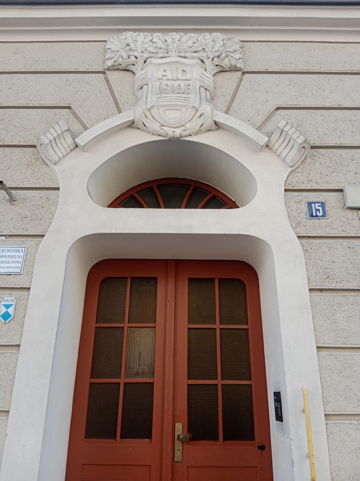 This is a frontal, eye-level photograph of an ornate doorway entrance to a building located at 15 Gdańska Street in Bydgoszcz, Poland. The entrance is framed by a white, stucco facade with horizontal grooved detailing. Above the doorway, a semi-circular window with red mullions is visible. A white decorative element is mounted above the window, featuring a crest with the inscription "A.D. 1903" and a stylized tree. To the left of the entrance, a blue and white sign reading "BYDGOSKA SPÓŁDZIELNIA MIESZKANIOWA ZARZAD I WSPÓŁWŁAŚCICIEL" is affixed to the wall, alongside a blue and white diamond-shaped emblem with the word "ZABYTEK". To the right of the doorway, the number "15" is displayed in blue on a white square plaque. The double doors are painted a vibrant orange-red and have a central brass doorknob and lock mechanism. A black intercom panel is mounted on the white wall to the right of the doors, and a yellow vertical pole is positioned to the far right of the frame. The overall scene depicts the entrance to a historic residential building.