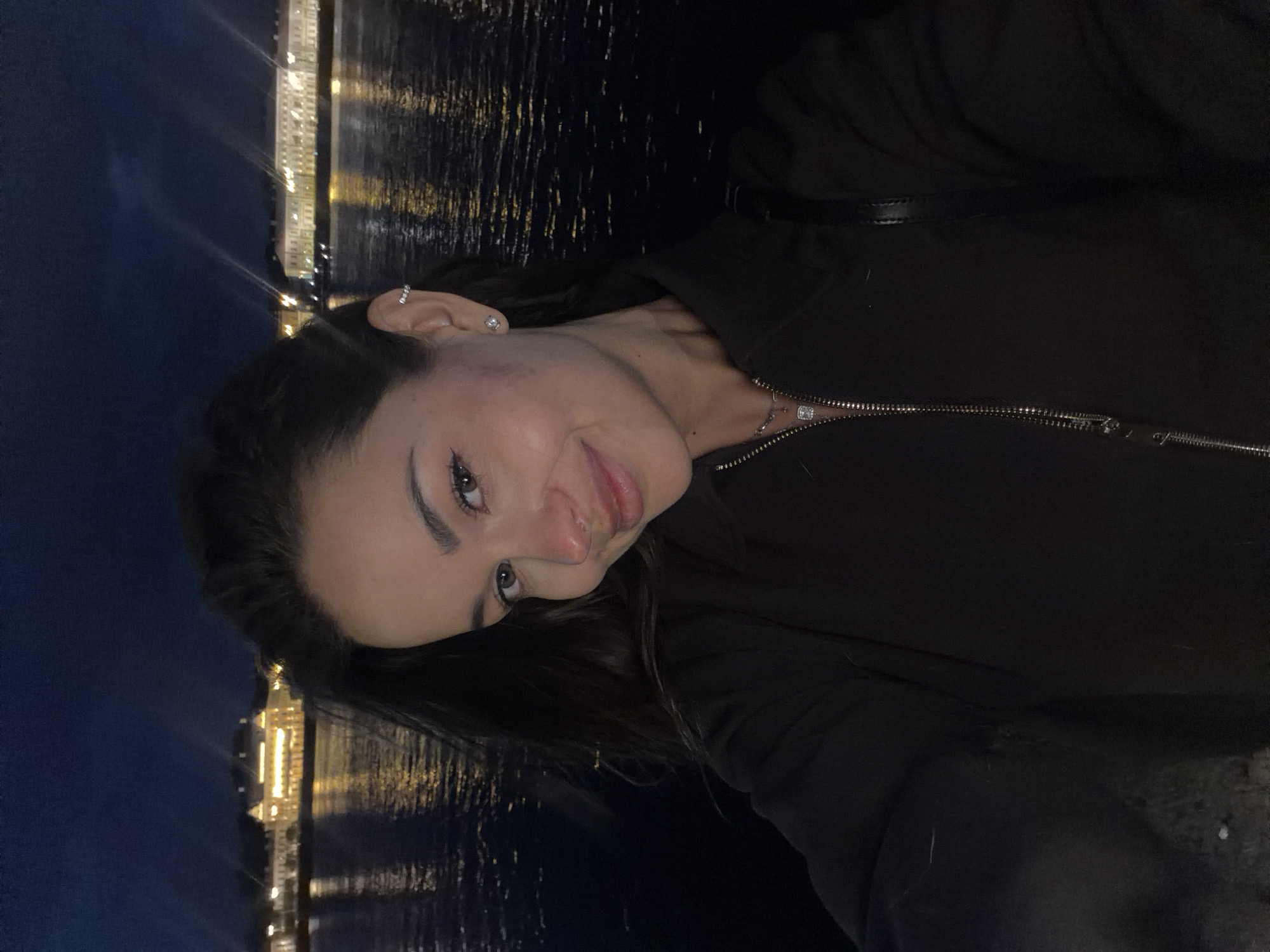 A young woman with dark hair and a light complexion takes a selfie at night by a body of water with buildings illuminated in the background. The woman is wearing a dark zippered jacket and a necklace. She has a piercing in her right ear and a nose ring. The background shows a waterfront with a row of historic buildings and their reflections in the dark water. The sky is dark blue, indicating it is nighttime. The overall scene suggests a location in Saint Petersburg, Russia, possibly along the Neva River embankment or a canal, given the architecture and waterfront setting.