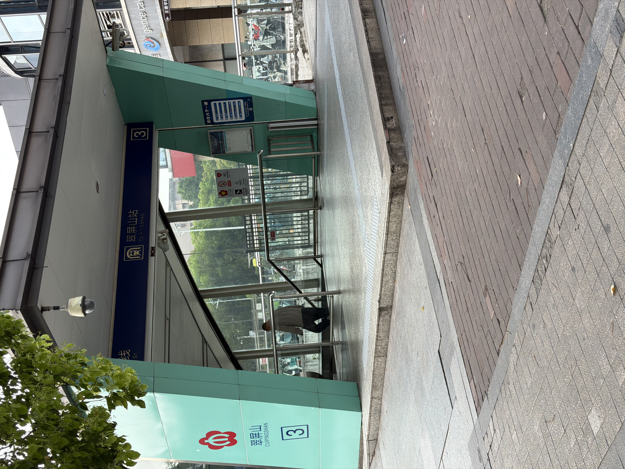 This image presents an outdoor view of a modern subway or metro station entrance, likely situated on a city street or sidewalk in Jiangning District, China. The scene is illuminated by bright daylight, suggesting a clear and dry day.
The central subject is the entrance to "翠屏山站" (Cuipingshan Station), with "CUIPINGSHAN" also displayed in Pinyin. The station entrance features a contemporary design with distinctive teal-colored panels and expansive glass sections, which reflect the surrounding urban environment, including trees and buildings. A prominent blue horizontal banner above the stairs clearly labels the station name and includes the number "3," reiterated on a square sign on the teal panel, likely indicating an exit number or line. A red, stylized flower or interconnected loop logo is visible on the lower teal panel.
A lone individual, wearing a light-colored jacket and dark pants, is captured descending the stairs into the station, engaging in the common activity of urban transit. Metal handrails line the staircase within the glass enclosure. A security camera is mounted above the entrance.
The ground comprises various paving materials, including grey concrete tiles, a brick-patterned path, and a grey tactile paving strip along the edge. Several pieces of text are visible on signs: "反作六个一律," a partially obscured "gemce cal," and "← 正德学院" (Zhengde Academy) in the background, indicating nearby landmarks or businesses. Additionally, a sign featuring "no smoking" and "no littering" symbols with the partially visible word "RED" is present. The overall impression is that of a functional and well-maintained urban transportation hub.