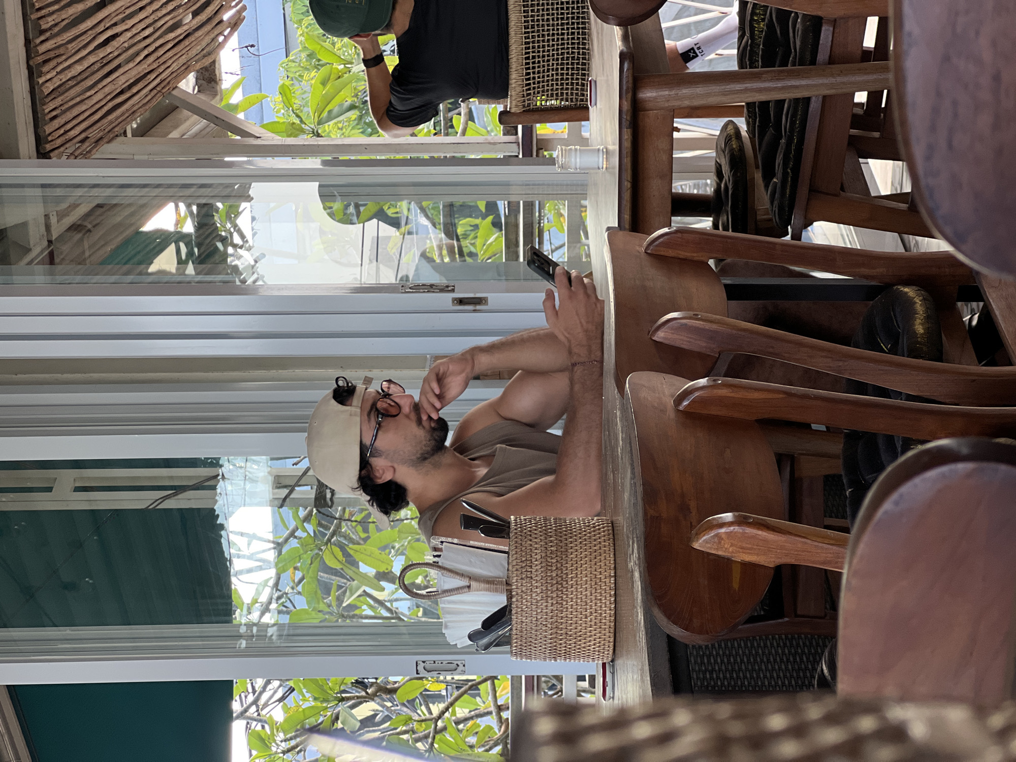 A man sits at a wooden table, indoors, looking at a phone in his hand. A window to his left shows an exterior of green trees. The man has on a hat and glasses, and a woven basket of napkins and silverware is in front of him. Chairs are stacked up in the foreground. The scene is in North Kuta, Indonesia.