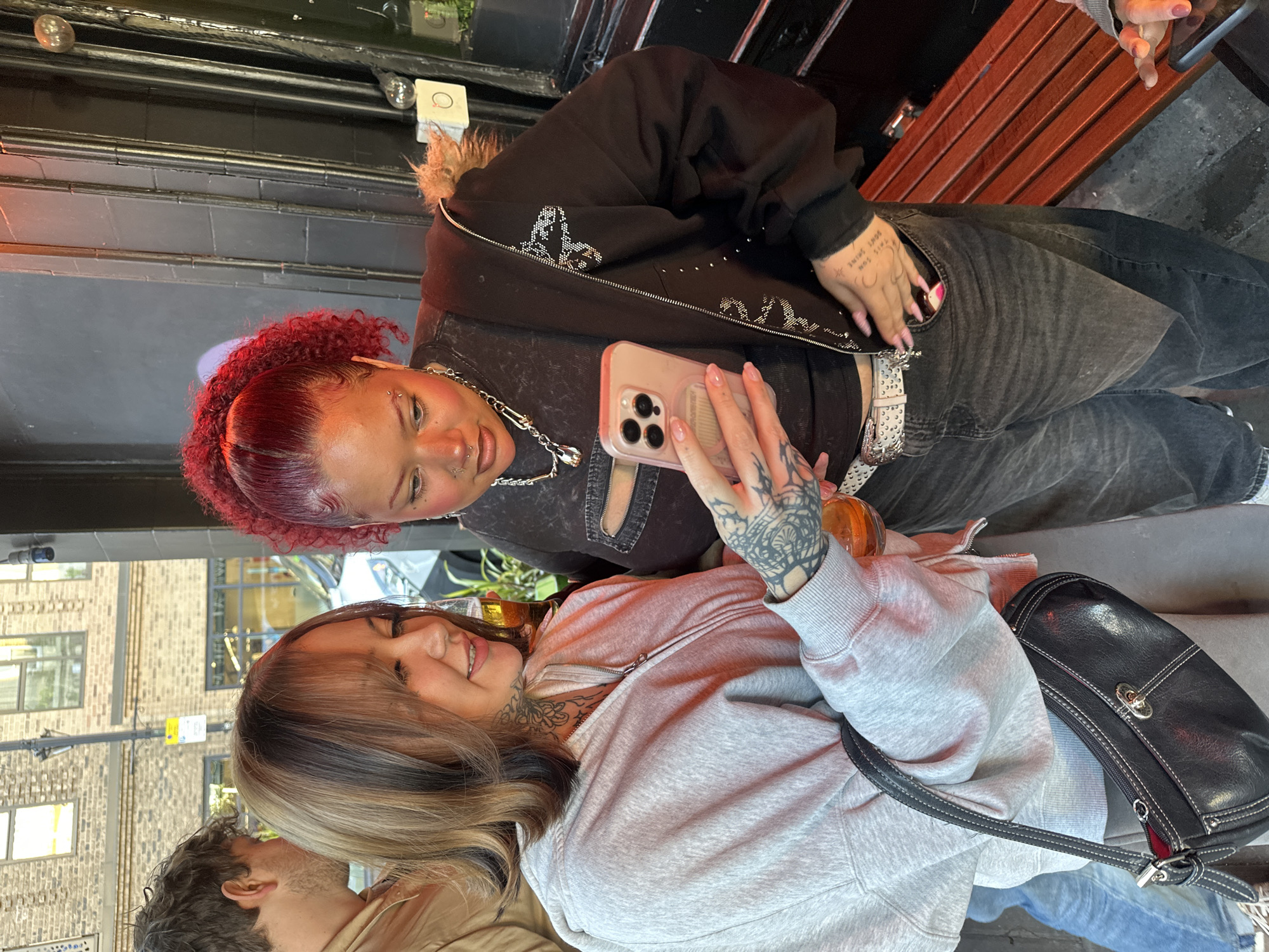 The image shows two women posing for a photo in an urban setting. The woman in the foreground has light brown hair with dark roots, a gray hoodie, and tattoos on her neck and arms. She is holding a mobile phone, presumably taking a selfie. The woman in the background has red hair, a black jacket with silver embellishments, and also has visible tattoos. Both women have visible piercings. They are seated outdoors, with a black leather handbag visible beside the woman taking the photo. Behind them are brick buildings, indicating an urban environment. Based on the architectural style and street signs, the scene is most likely set in the city of London, United Kingdom.