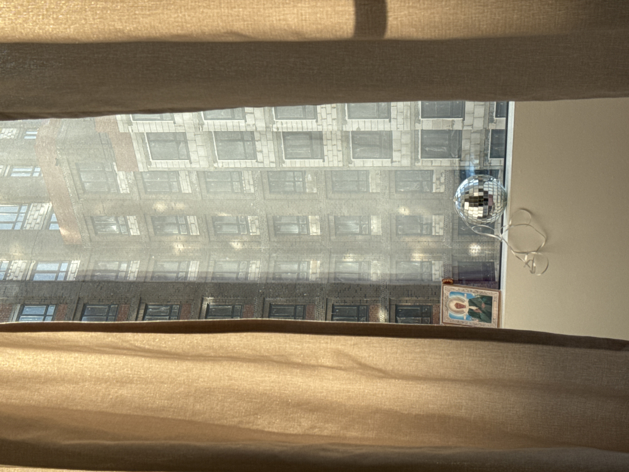 The image shows a partial view from a window covered by a light-colored curtain. Through the opening in the curtains, a building across the street is visible, featuring a brick facade and a grid of windows. Inside the room, adjacent to the window, there's a disco ball hanging with a wire. A small framed picture is situated next to the window as well. There are no visible people, actions, or interactions. The location, Люк, Russia, is not ascertainable from the image.