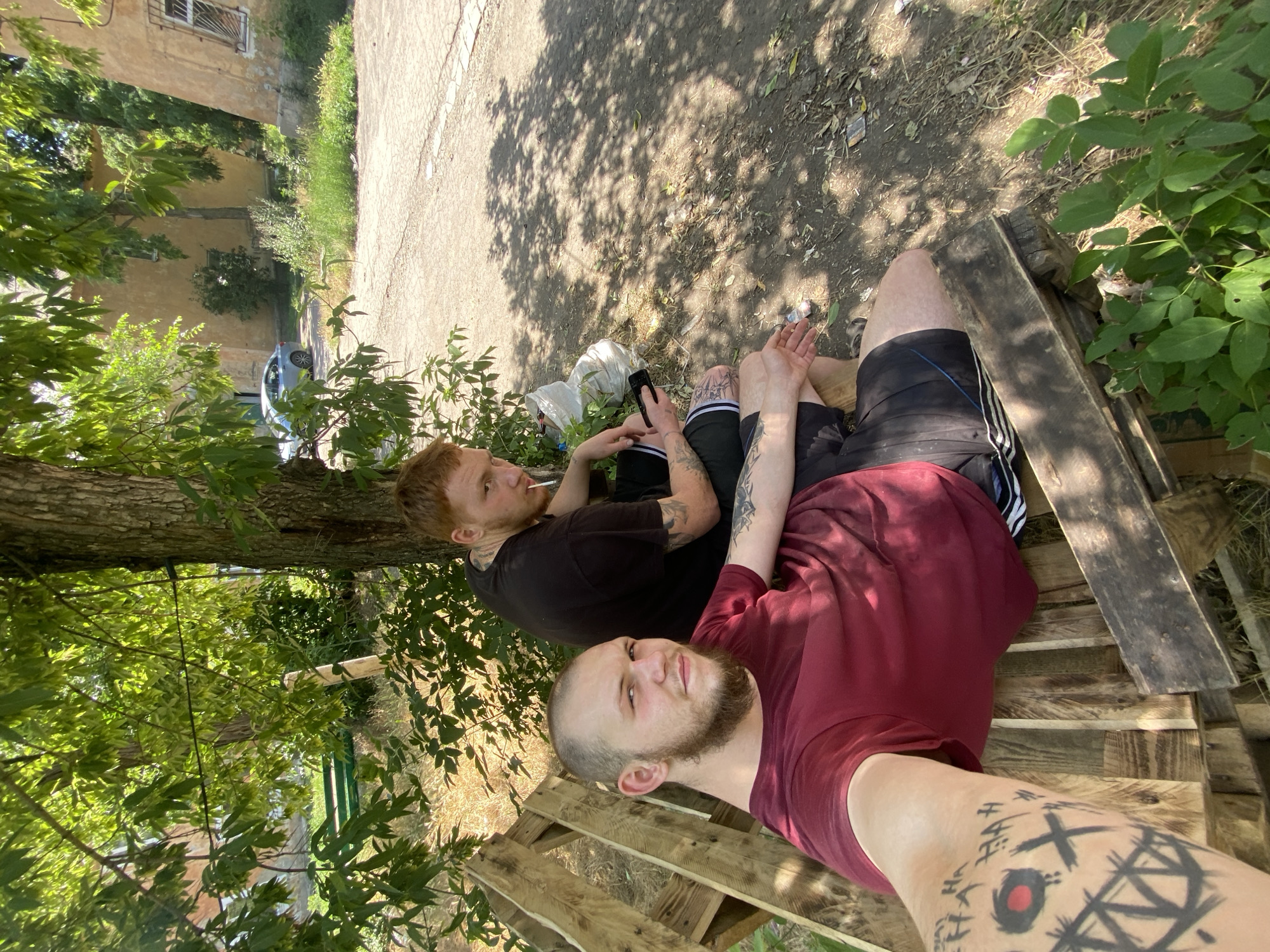 Two men are sitting on a wooden bench outdoors. The man in the foreground is wearing a maroon t-shirt and black shorts. He has a beard and tattoos on his left arm. The man in the background is wearing a black t-shirt and black shorts. He has red hair and is holding a cigarette in his mouth. He is also holding a cell phone in his left hand. The bench is made of wooden pallets and is situated under a tree. In the background, there are buildings and a car. The ground is covered with dirt and leaves. The lighting suggests it is daytime.