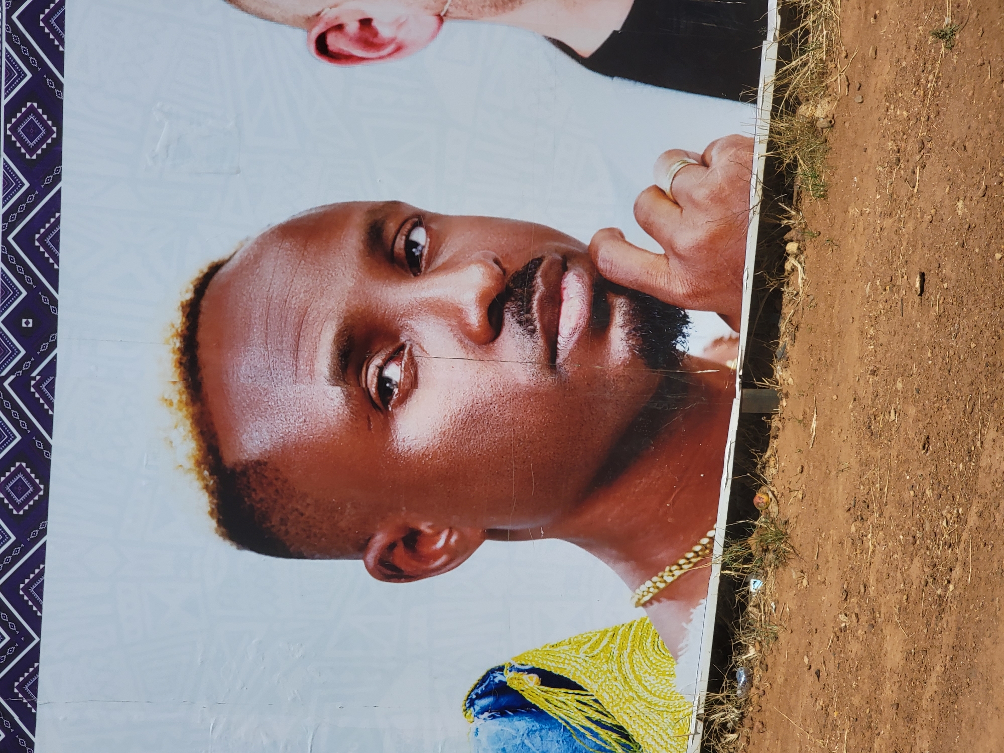 The image shows a partially deteriorated promotional poster leaning against the ground in Jos, Nigeria. The ground is reddish-brown dirt with scattered grass. The poster features the close-up face of a dark-skinned man with a light-colored hairstyle and a short beard. He wears a gold chain and a ring on his finger. In the upper part of the poster, the top of a light-skinned person's head is visible. Part of a blue and yellow patterned design is at the bottom of the poster. The left side of the poster has a patterned purple and white border.