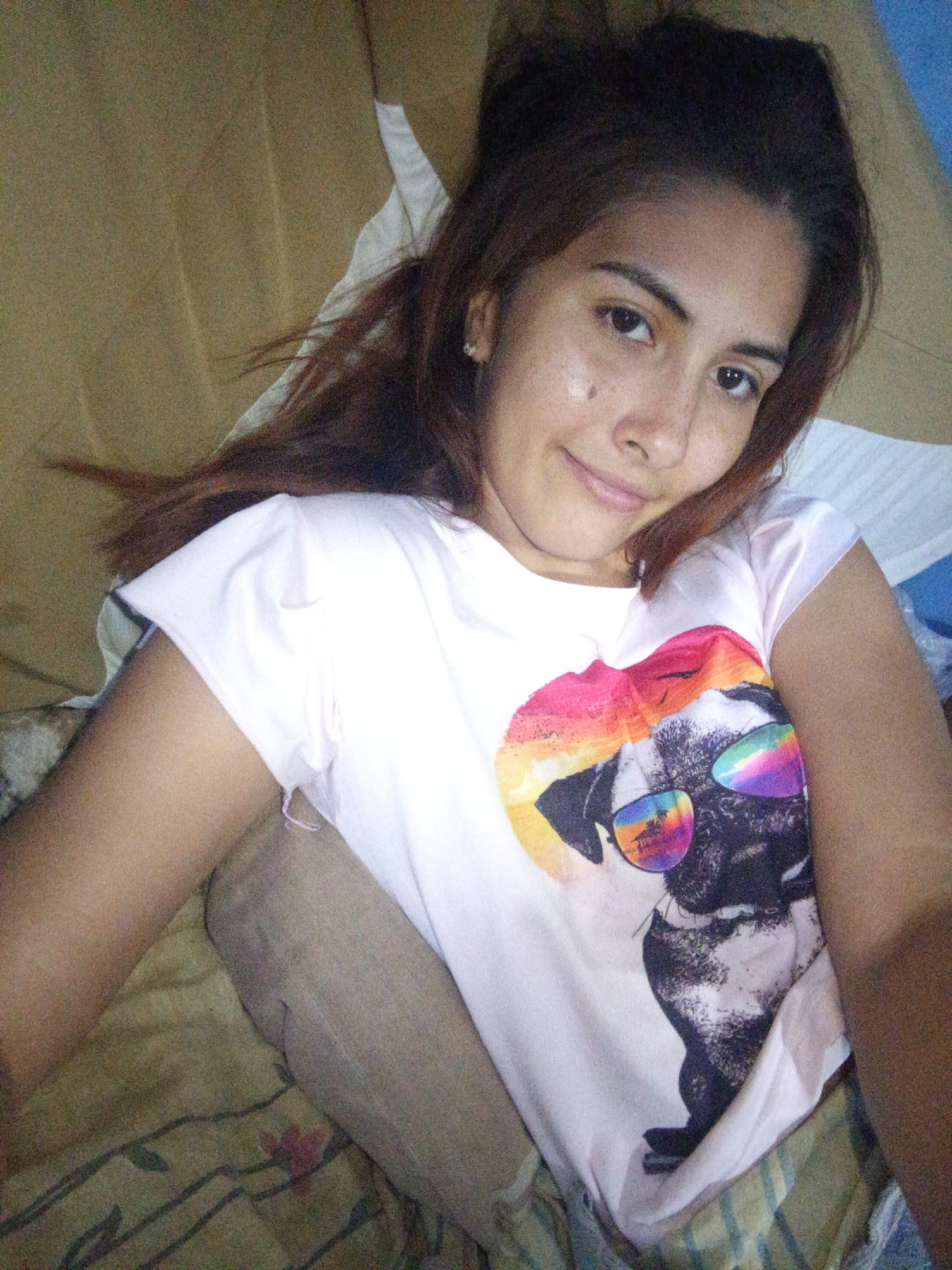 A young woman with long, dark brown hair is lying down on a bed. She is wearing a white t-shirt with a graphic of a pug wearing sunglasses. The background appears to be a bedroom or living room with beige and green fabrics. The lighting is soft and natural. There are no visible landmarks that would indicate a specific location in Juan Griego, Venezuela.