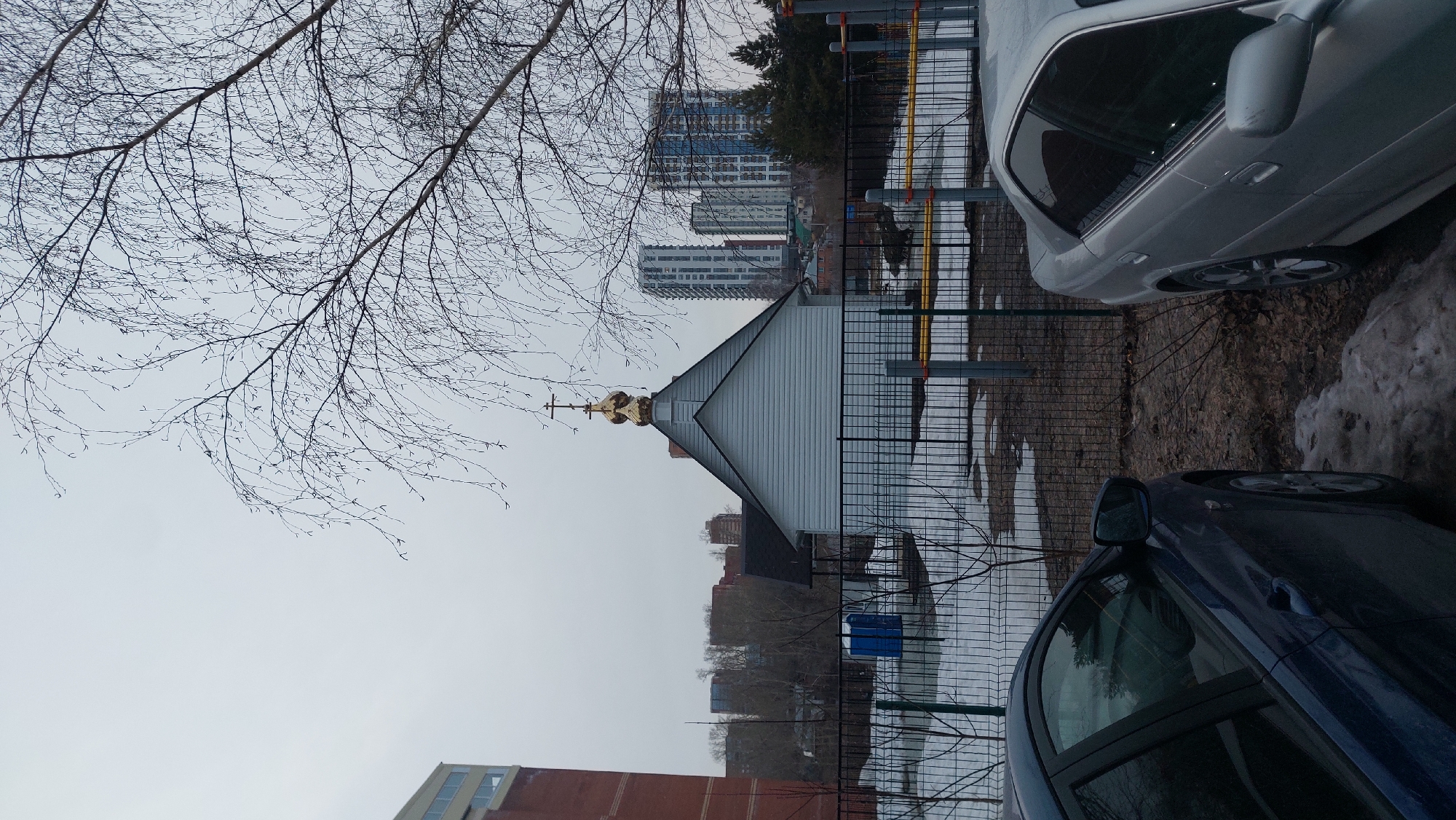 The image shows a small church with a golden dome and cross in Novosibirsk, Russia. The church is located near modern high-rise apartment buildings. A bare tree branch extends into the frame from the upper left. The sky is overcast and grey. There is a metal fence separating the church from a patch of ground with melting snow. Two cars, a silver one and a dark blue one, are parked nearby.