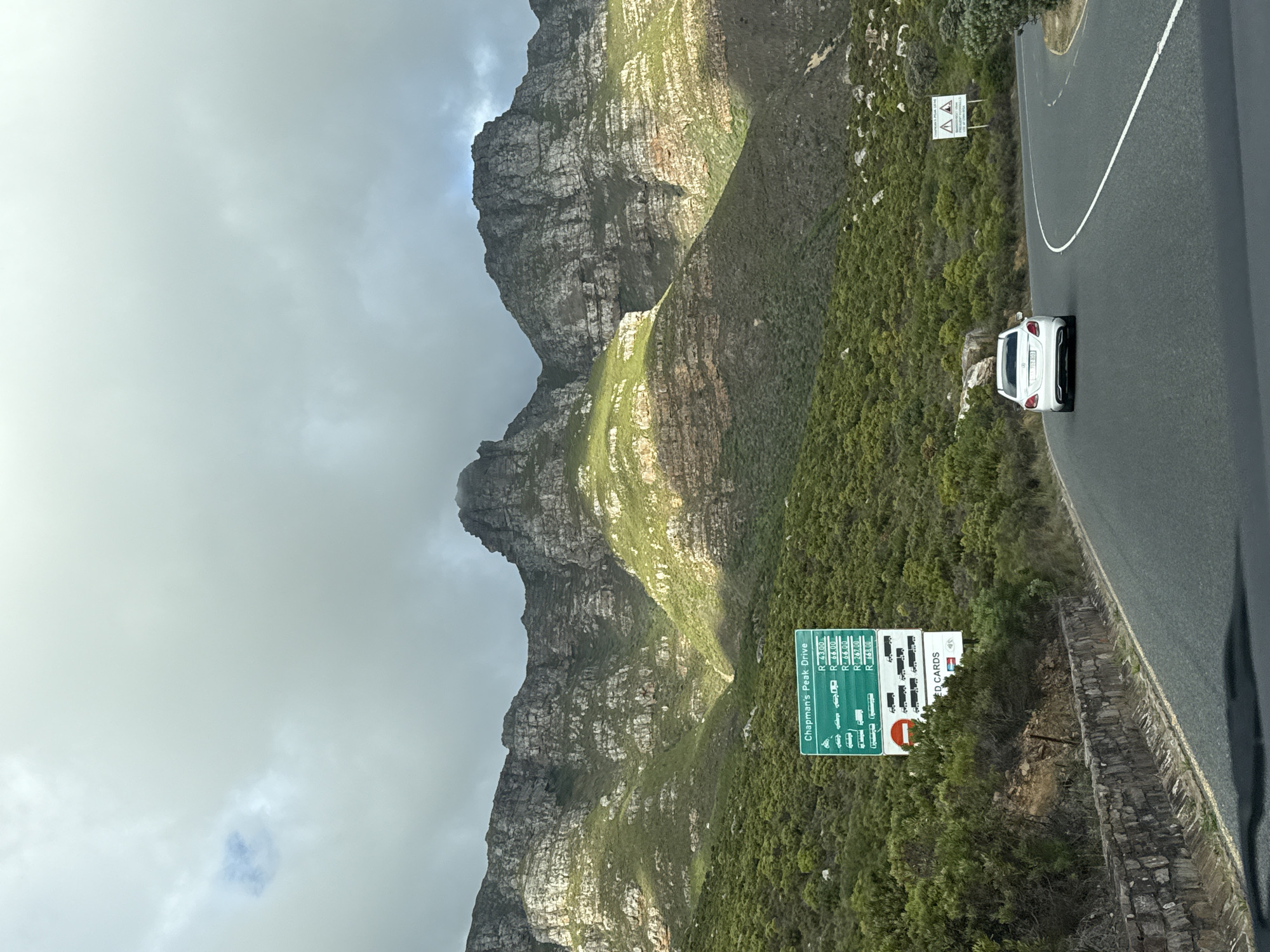 The image shows a mountain landscape, likely in Hout Bay, South Africa, given the "Chapman's Peak Drive" toll sign. The mountain is rugged, with visible rock formations and some green vegetation. A white car is on the road running along the mountainside. There are road signs present. The sky is overcast with clouds.