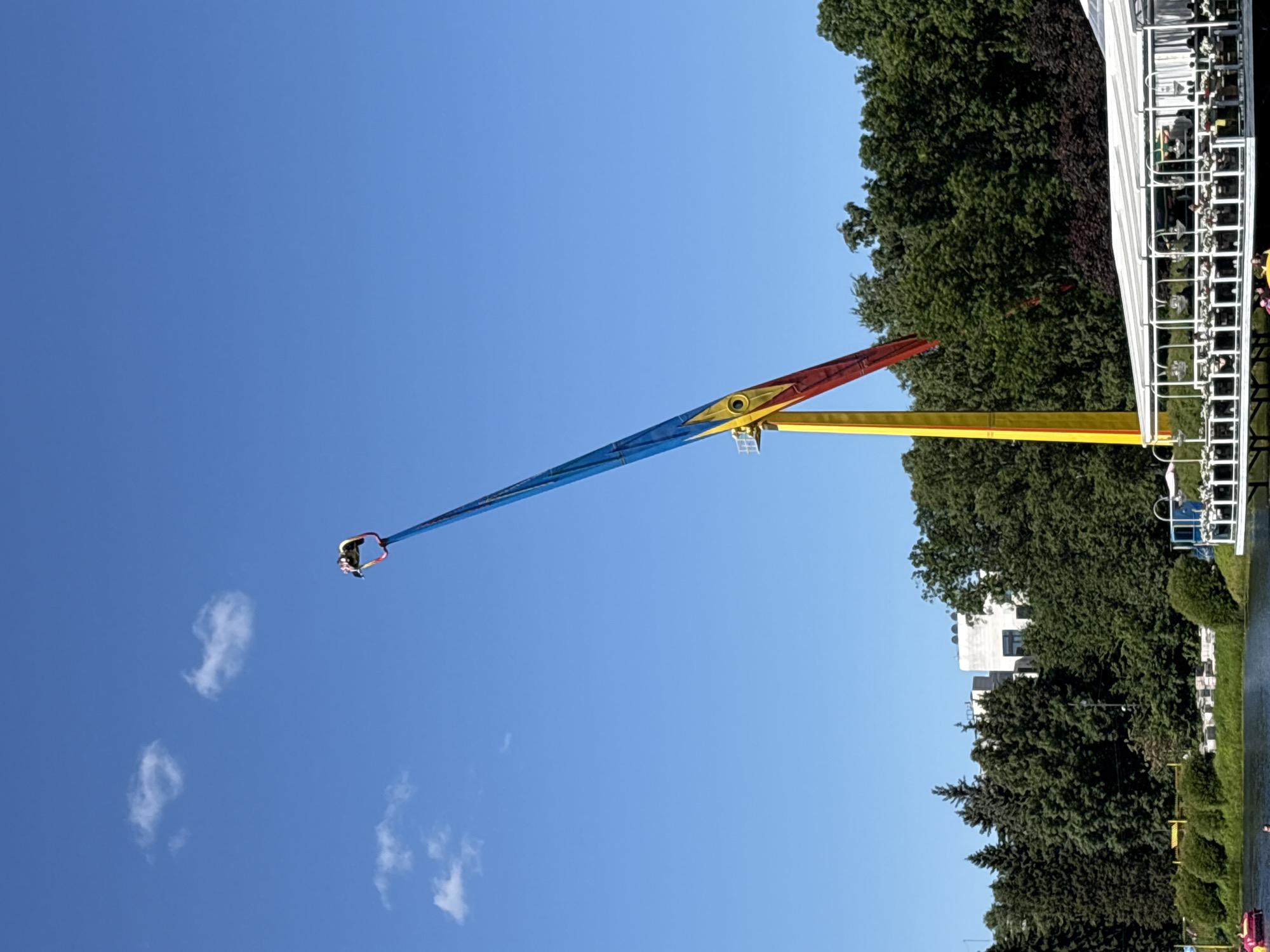 This media file captures a thrilling amusement park ride against a clear blue sky, with lush green trees and a white building forming the backdrop. The central subject is a large, colorful ride that appears to be a pendulum or swing attraction. It features a long, slender arm with sections colored blue, yellow, and red, adorned with yellow starbursts. At the very end of the arm, a single carriage with a person seated is visible, suspended high in the air, suggesting the ride is in motion or at its peak.
The scene is set in Saint Petersburg, Russia, under what appears to be pleasant weather with a bright, cloudless sky, indicating it is likely daytime, possibly late morning or afternoon. The presence of dense foliage and a modern building hints at an urban or suburban park setting. The atmosphere seems energetic and exciting, given the nature of the ride and the clear skies. The image is framed such that the ride extends diagonally across the frame, emphasizing its height and reach. There is no visible text in the media file.