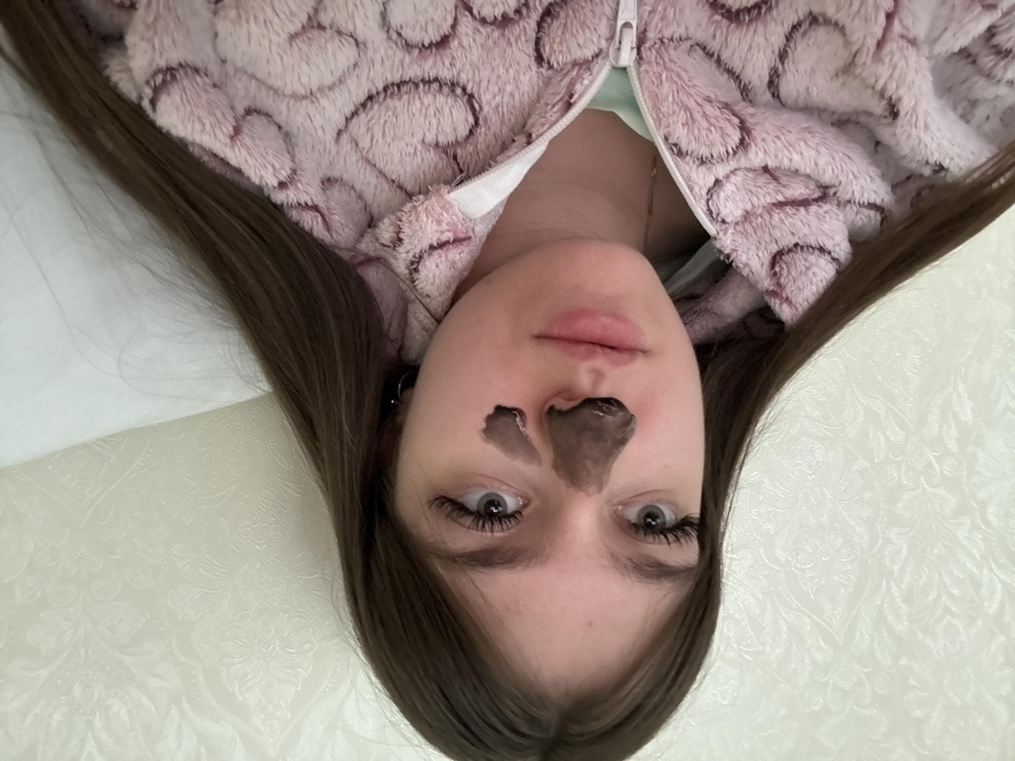 A young woman with long, dark brown hair is captured in a close-up, top-down shot, appearing to lie down. Her head is oriented towards the top of the frame, and she gazes directly at the viewer with wide, grey or blue-grey eyes, enhanced by long, dark eyelashes.
She is wearing a plush, light pink hooded zip-up jacket or loungewear, which features a repeating pattern of darker pink, abstract, swirling shapes. A light green garment is visible underneath the partially unzipped jacket. A dark-colored (likely black or charcoal) peel-off mask is applied to her nose and the area between her eyebrows. Her lips are naturally pink and slightly parted. A small, metallic earring is discernible in her left ear.
Her dark hair frames her face, fanning out onto a light-colored, smooth surface beneath her. The background consists of a light-toned, possibly off-white, wall adorned with an embossed, intricate pattern reminiscent of floral or damask designs. The lighting is soft and uniform throughout the image. No text is visible.