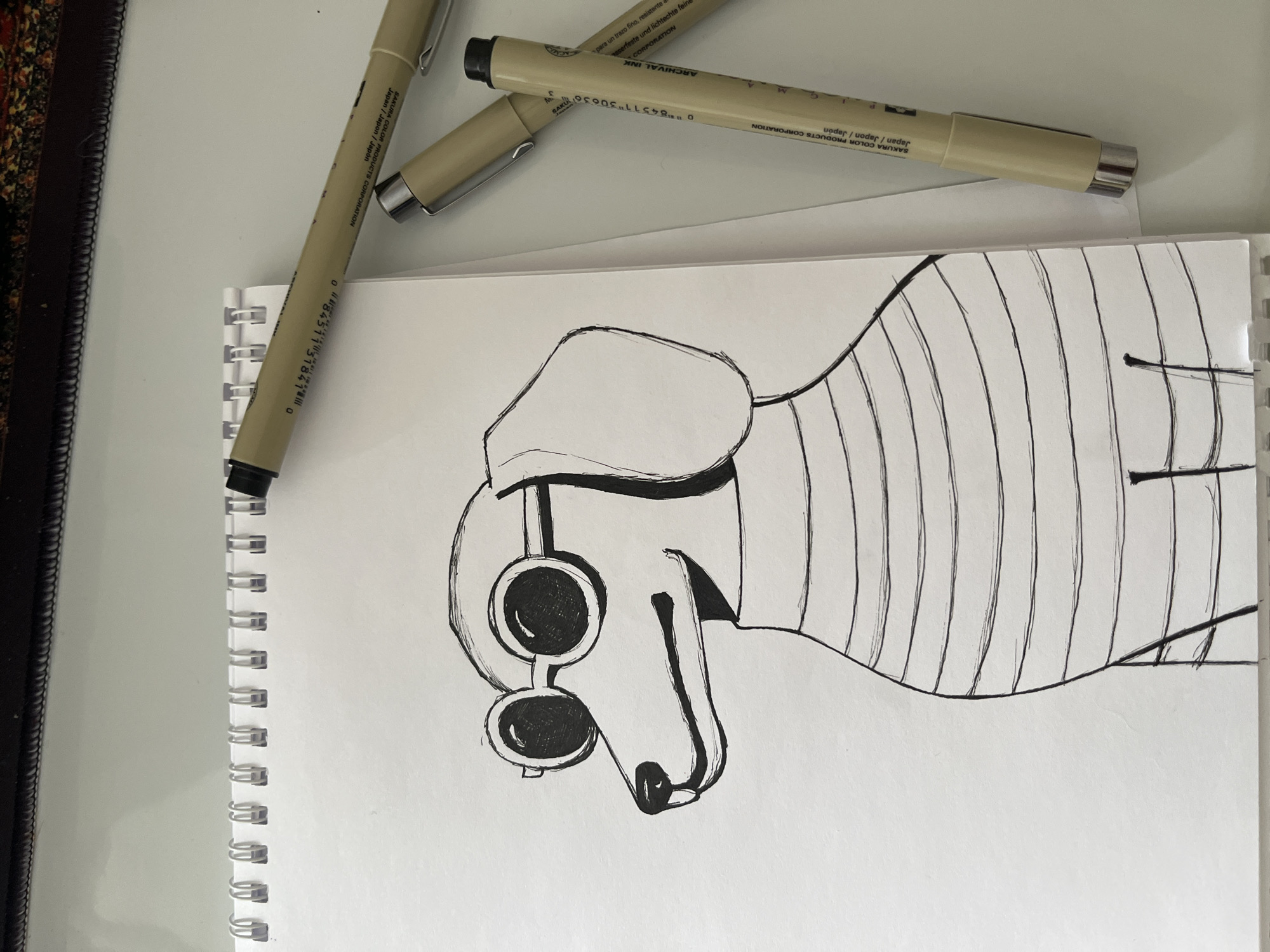 The image is a close-up shot of an open sketchbook on a white surface, likely a table. Three light brown Sakura Pigma Micron pens are arranged near the top of the sketchbook. The sketchbook contains a drawing in black ink. The drawing depicts a stylized figure with a hat and goggles, possibly a cartoon character or a stylized animal. The figure appears to have a long neck or body section with vertical lines drawn across it. The sketchbook is spiral-bound on its left side. A dark-colored object, potentially a frame or part of a piece of furniture, is visible along the extreme left edge of the image. The environment suggests an indoor setting, possibly an artist's workspace. There is no indication of Novosibirsk, Russia in the image.