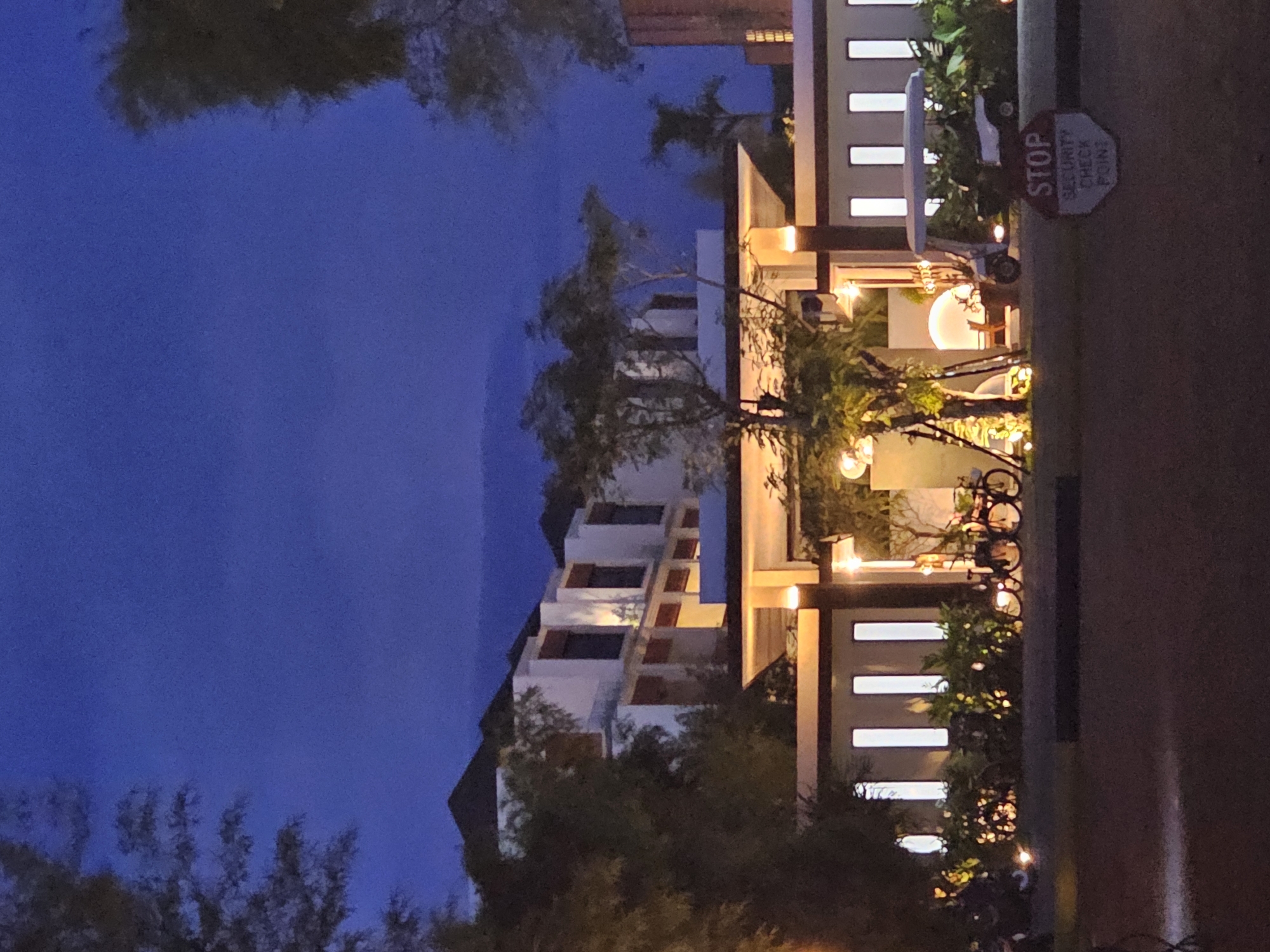 The image, taken at night, shows a building in Denpasar, Indonesia. The building is white with dark brown accents and has multiple levels. The windows are lit up. A "STOP SECURITY CHECK POINT" sign is visible on the right side of the image, near a golf cart. There are trees and bushes surrounding the building, some of which are lit up with string lights. The sky is dark blue.
