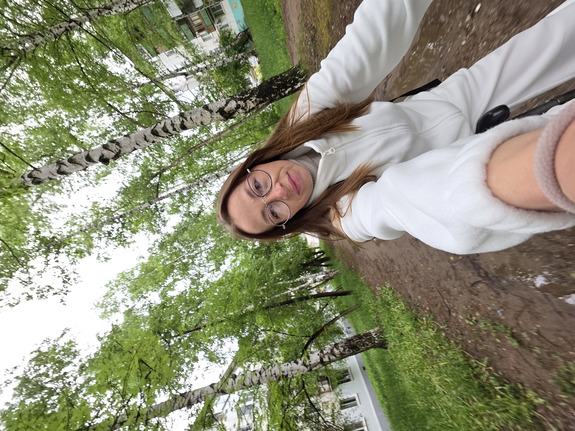 A selfie captures a woman in a white fleece jacket and pants, wearing round glasses, and a pink beaded bracelet on her left wrist. The background reveals a city park setting, identifiable by birch trees with their characteristic black and white bark, lush green foliage, and a muddy path. Buildings are visible in the background, suggesting an urban environment. Given the prompt's specification, the location is likely Chaikovsky, Russia. The woman is positioned on what appears to be a bicycle, implied by the presence of a black handlebar in the frame. She is directly facing the camera, capturing the selfie from a low angle.