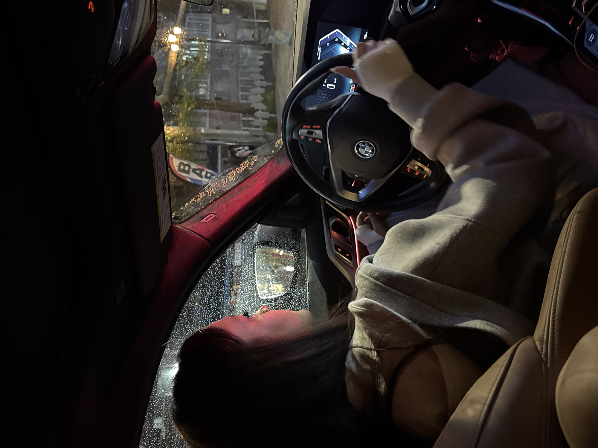 A woman is seated in the driver's seat of a BMW, her hands on the steering wheel. The car is likely stationary, as the woman is gazing out the rain-covered windshield. The scene appears to be at night in Yerevan, Armenia. The view through the windshield shows a blurred urban environment with streetlights and signs. Based on a partial sign visible through the windshield that reads "B...", the car is located near a BAB Döner.