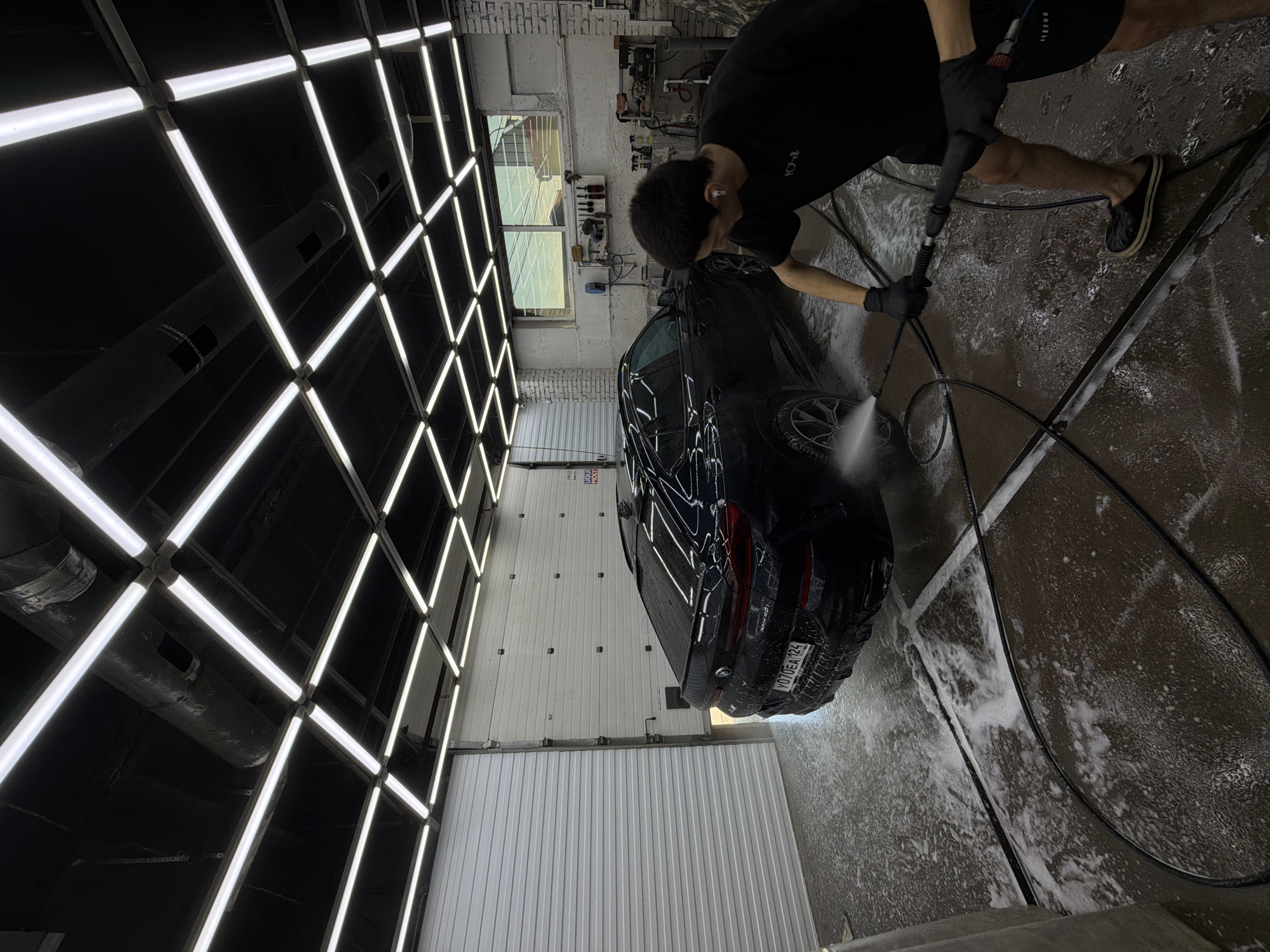 A man in a black t-shirt and shorts, wearing black gloves and black slip-on shoes, is power washing a black BMW X6 in a car wash bay in Krasnoyarsk, Russia. The car is partially covered in soap suds and water. The car wash bay has a white roll-up door on the left, white brick walls, and a grid of fluorescent lights on the ceiling. Shelves with cleaning supplies and tools are mounted on the back wall. The floor is wet and covered in soap. The license plate of the car is partially visible and reads "Y070EA 124".