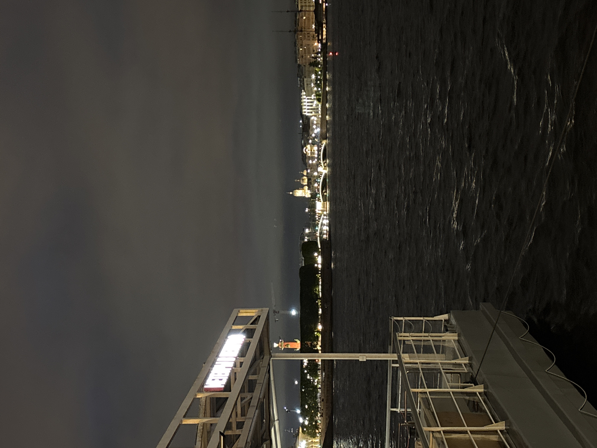 The image shows a nighttime view of the city of Saint Petersburg, Russia, taken from a pier or dock. The Neva River stretches horizontally across the right side of the image. The city skyline is illuminated with lights, featuring landmarks such as bridges and buildings with architectural details. The sky is overcast. In the foreground, there is a structure with a sign and metal framework, likely part of the pier or dock. A white railing is also visible on the right side of the image. There are no people visible in the image.