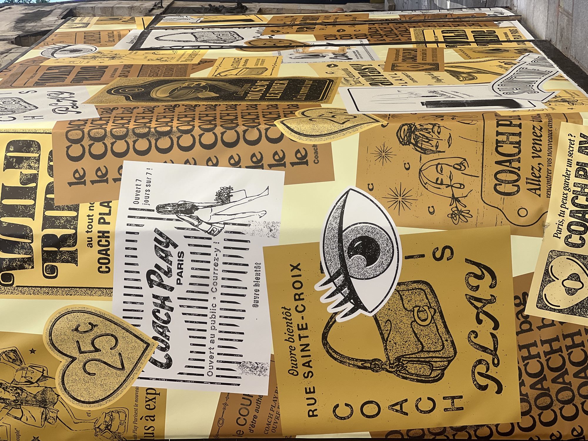 The image depicts a wall covered in a collage of posters, predominantly in shades of yellow and black, which appear to be advertisements or promotional materials for the brand COACH PLAY. The posters feature a graphic, retro-inspired aesthetic with illustrations of handbags, stylized eyes, fashion figures, and text. Some posters are in French, with phrases like "Ouvre bientôt" (Opening soon), "Au tout nouveau COACH PLAY Paris," and "RUE SAINTE-CROIX." The collage is dense, with posters overlapping and some peeling at the edges, suggesting an outdoor urban environment. The backdrop of the wall appears to be a textured, light-colored surface, possibly stucco or stone, consistent with the architecture of Paris. One poster clearly indicates "PARIS" in its text, and the presence of French language further supports the Paris location. The style of the wall and the overall presentation suggest this is a temporary street art installation or a facade decoration for a store or event in the city.