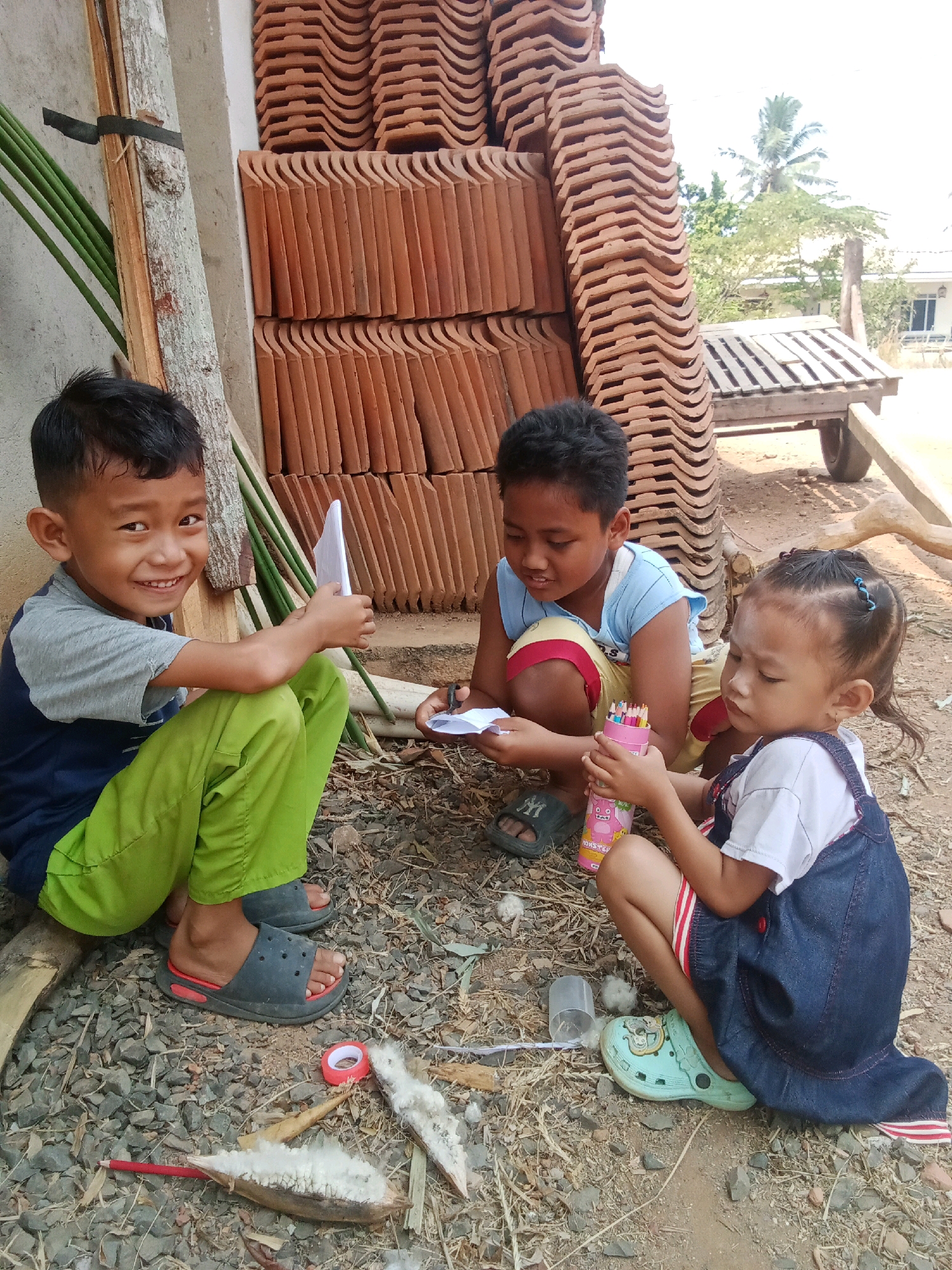 In an outdoor setting, possibly in the city of Unknown, Indonesia, three children are seated on the ground amidst debris and building materials. To the left, a young boy in a blue and gray shirt and bright green pants smiles at the camera, holding a white sheet of paper. Next to him, another boy in a blue tank top and red-trimmed shorts is also looking down at a piece of paper, appearing to be drawing or writing. A young girl in a denim overall dress and white shirt is crouched to the right, holding a pink cylinder filled with colored pencils. The background features a stack of reddish-orange roof tiles, some green bamboo stalks leaning against a rough-textured wall, and a wooden cart with wheels. The ground is covered with gravel, dirt, and various natural elements such as seed pods and a red spool of tape. The overall scene suggests a moment of casual activity and play outdoors.