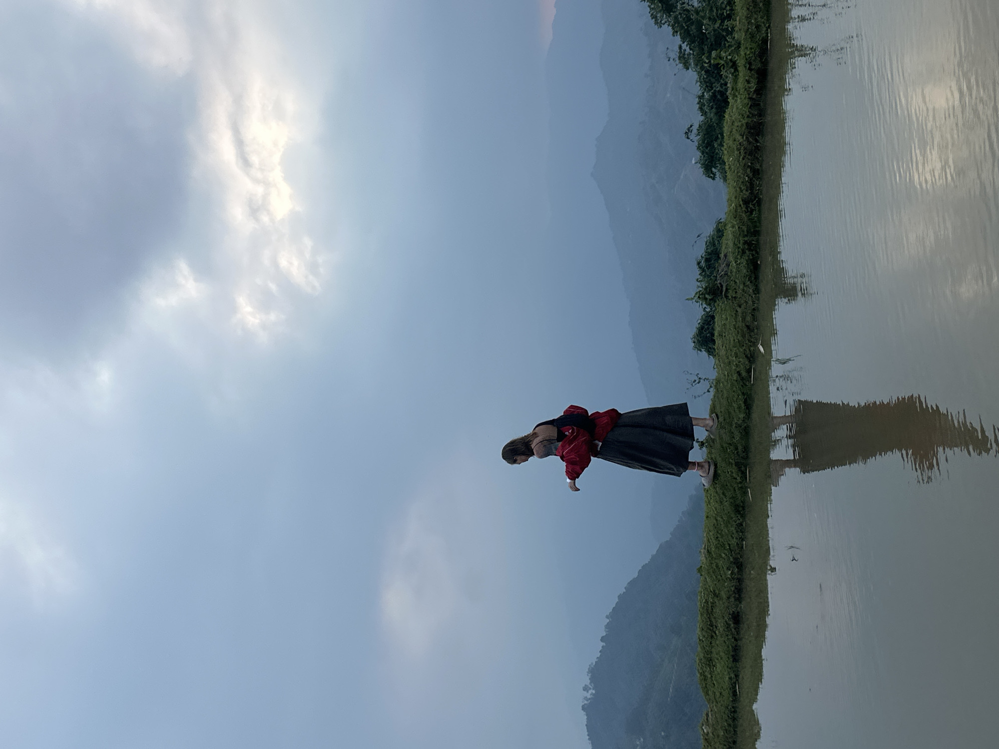 A full-shot image, originally in portrait orientation but rotated 90 degrees counter-clockwise, depicts a person standing on the edge of a body of water with a mountainous backdrop under a cloudy sky.
The central subject is a person, likely a woman, seen from the side, standing on the grassy bank of a calm water body. She is wearing a red jacket over a grey top, a dark skirt, and light-colored shoes. Her arms are slightly bent, and she appears to be looking downwards, possibly at the water or something on the bank. Her reflection is visible in the water, though slightly distorted by ripples.
The scene is dominated by a vast sky that transitions from a lighter, cloud-filled area on the left to a slightly darker, hazy blue on the right. Several fluffy, white clouds are visible, particularly in the upper left. In the background, a series of mountains rise, appearing misty and layered, suggesting distance and elevation.
A strip of lush green vegetation, possibly grass and small bushes, forms the immediate bank between the person and the water. The water itself is relatively still, reflecting the sky and the distant mountains with a soft, diffused quality. Ripples on the water surface create subtle texture.
The lighting suggests it is daytime, possibly late afternoon or early evening, given the soft, somewhat diffused light and the atmospheric haze over the mountains. The overall mood is tranquil and natural. No visible text is present in the image. The location is Nậm Dịch Commune, Vietnam, which aligns with the natural landscape and mountainous features.