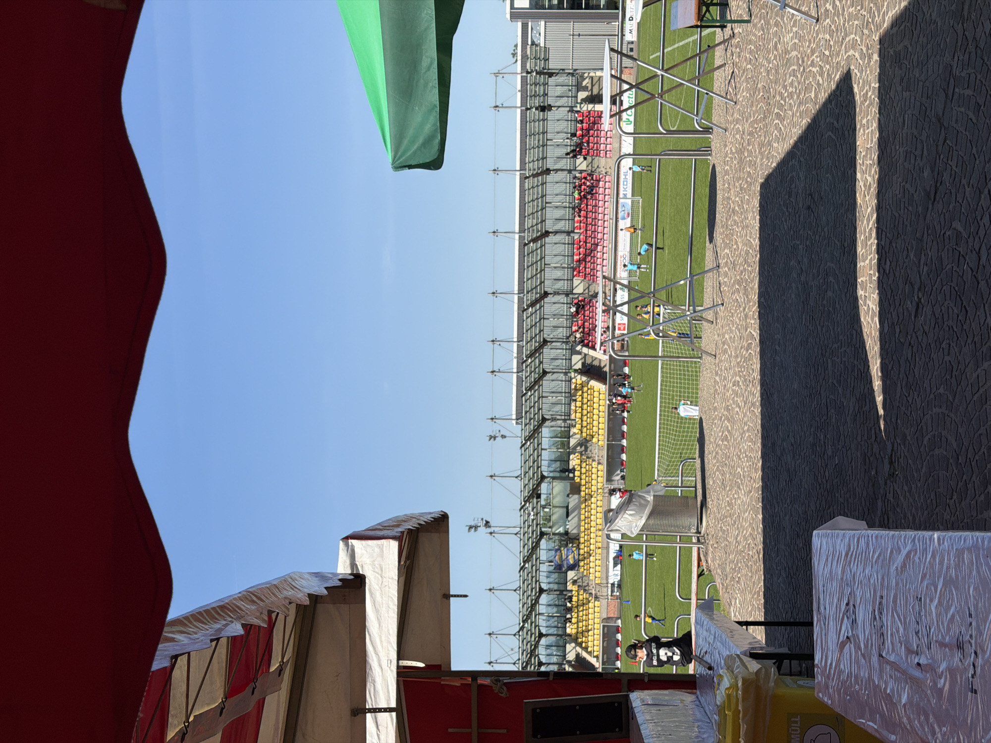 The image shows a view of a soccer field with red and yellow seating visible in the stadium. The soccer field appears to be located in Fentange, Luxembourg. The perspective is from behind a vendor's area, with a red awning on the left, and a green awning visible above the field. A cobblestone walkway is on the right, in shadow. There are people visible on the field, and it appears to be a game or practice in progress. There is a garbage can and what looks like packaged items in the foreground. The sky is blue and clear.