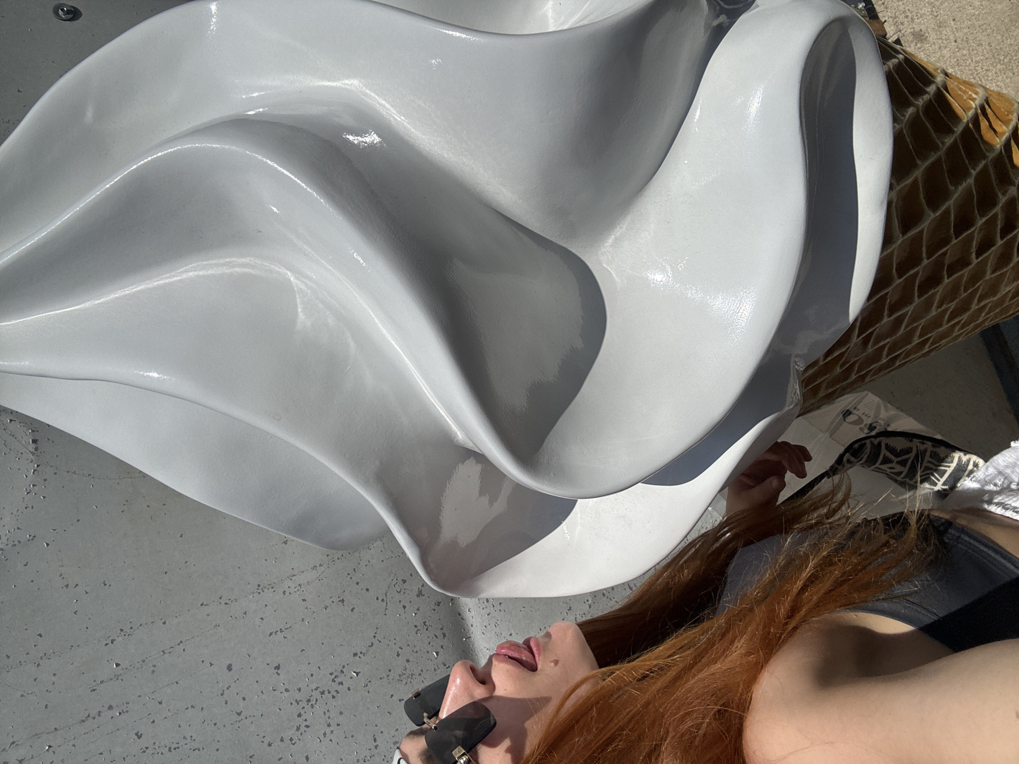 In Budva, Montenegro, a woman with long red hair and sunglasses playfully sticks her tongue out towards a large, white, sculpted soft-serve ice cream cone. She is positioned on the left side of the frame, with her head tilted and her gaze directed at the sculpture. The ice cream cone, depicted in a waffle cone, dominates the right and center portions of the image. The background consists of a textured concrete wall on the left and a sunlit ground surface on the right. The lighting suggests a bright, sunny day, casting distinct shadows. The woman is wearing a dark top and appears to be near a shopping bag.