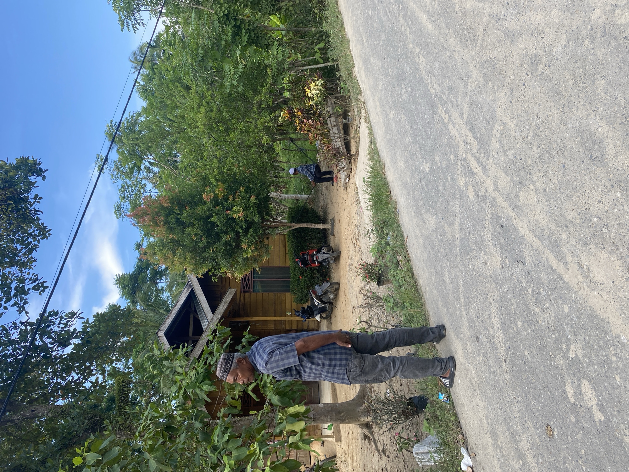 A man stands in Gampongaceh, Indonesia, next to a rural road. He wears a patterned shirt, dark pants, and a cap. Behind him is a small wooden house and two parked motorbikes. Trees and vegetation surround the buildings and the road. Another individual stands further down the road, near a small structure. The sky above is blue with some clouds.