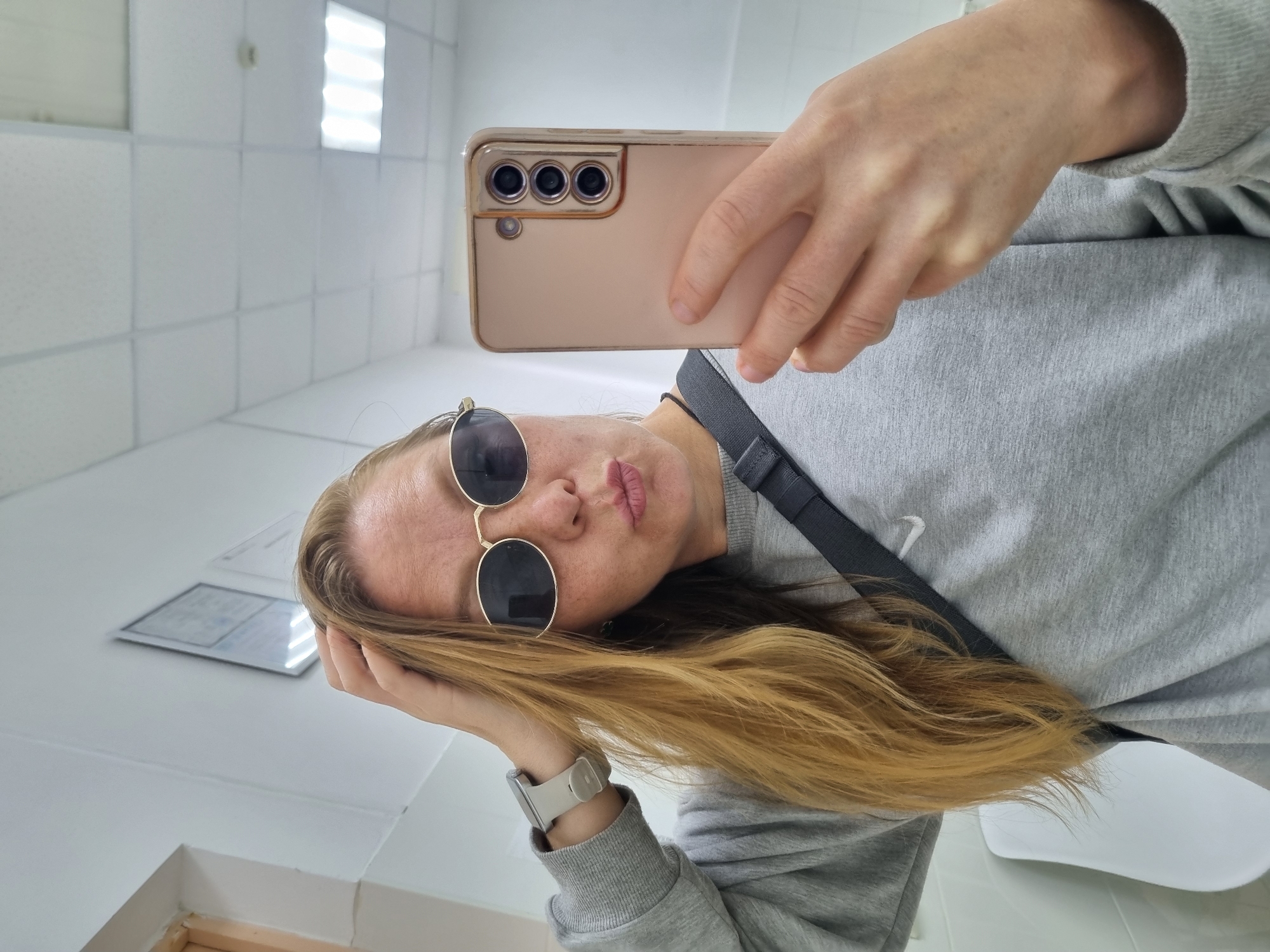 A woman with blonde hair and sunglasses is taking a selfie in a bathroom. She is wearing a gray sweatshirt and has a black strap across her chest. The bathroom has white tiled walls and a fluorescent light fixture on the ceiling. The location is identified as Chaikovsky, Russia.