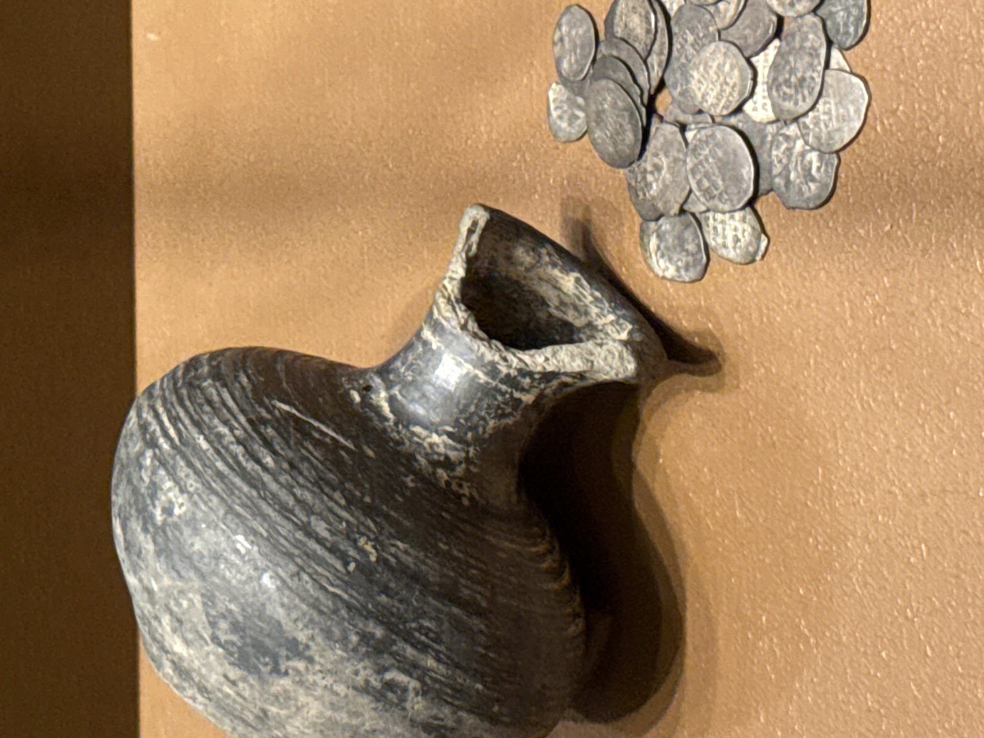 The image shows a grey, ancient-looking pitcher and a small pile of silver coins. The items are displayed on a brown surface or backdrop, possibly within a museum exhibit. The lighting casts shadows, highlighting the texture and shape of the pitcher and coins. The scene is likely taken at the State Historical Museum in Moscow, Russia, where many ancient artefacts and treasures are displayed.