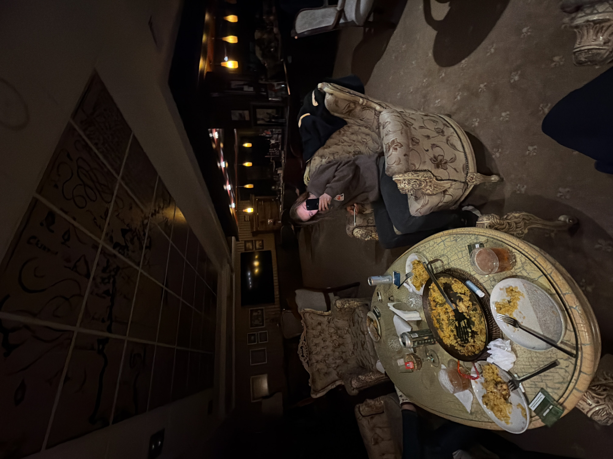 The image shows the interior of a restaurant in what is possibly the city of Silistra, Bulgaria. A woman is seated in an ornate chair, looking at a phone. A table laden with plates, cutlery, and food is in front of her. The food appears to be paella or a similar rice dish. There are also beverage containers and what appears to be a pack of cigarettes on the table. The restaurant has ornate decor, including patterned chairs and a large mural on the wall. Further in the background, there are other tables and chairs. A television is mounted on the wall.