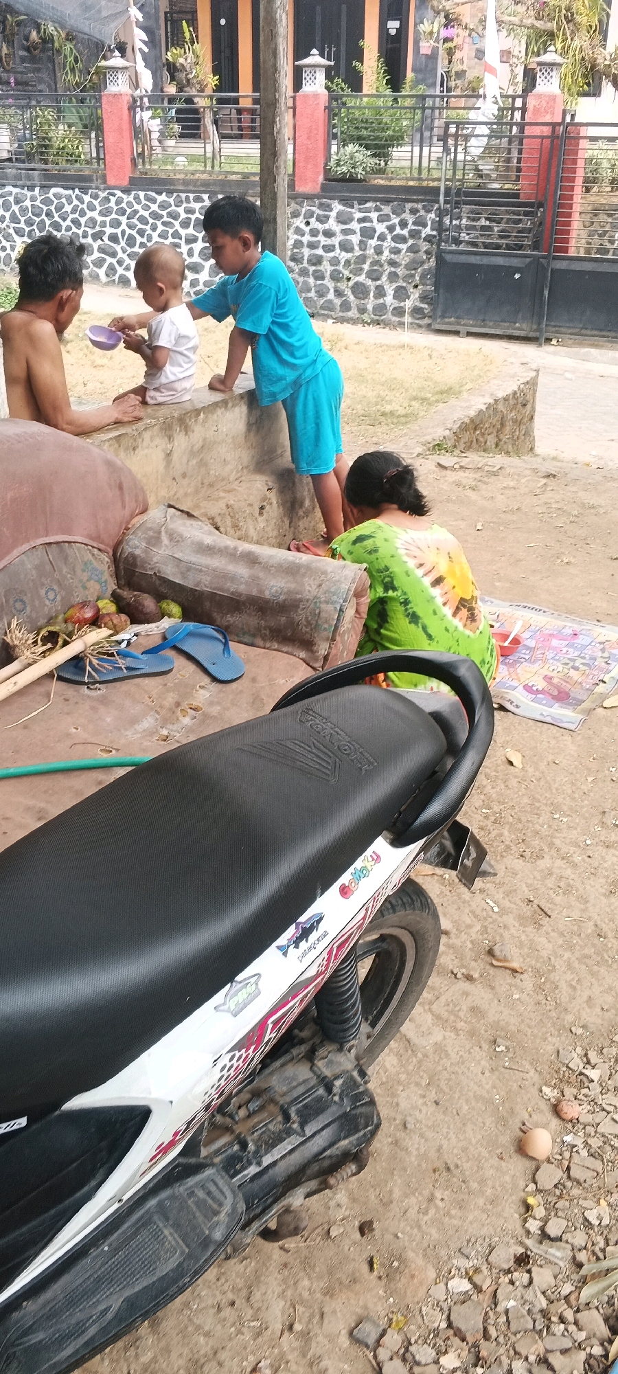 In Penanggungan, Indonesia, a family is gathered outside a home. A man, partially undressed, sits on a low concrete wall. Beside him, a baby in a white shirt and light-colored shorts is being fed by an older child in a blue t-shirt and shorts. The older child is holding a purple bowl and spoon, interacting with the baby. Another individual, a woman wearing a green tie-dye shirt, sits on the ground facing away from the camera, looking at a mat spread out on the ground. The mat has colorful patterns and appears to have some items on it. Behind the people, there is a house with orange accents and a black metal fence. The ground is dusty and dirt-covered, with scattered stones and debris. A motorcycle with a black seat and white bodywork is parked in the foreground. On the ground near the motorcycle, there are blue flip-flops, some fruits, and dried plant material. The scene suggests a casual, outdoor family moment in a residential area.