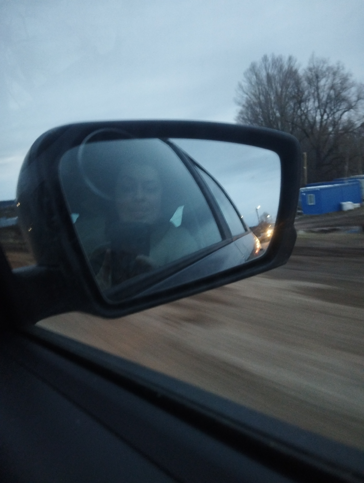 The image shows a side view mirror of a moving vehicle. The mirror's reflection shows a person, likely the driver, and the outside environment. The outside environment includes trees without leaves, a blue building, and a road, likely located in a rural or suburban area near Votkinsk, Russia. There are other lights from vehicles visible. The sky is overcast, suggesting it is either evening or early morning. The image is slightly blurred, indicating motion. The interior of the vehicle is visible in the lower left.