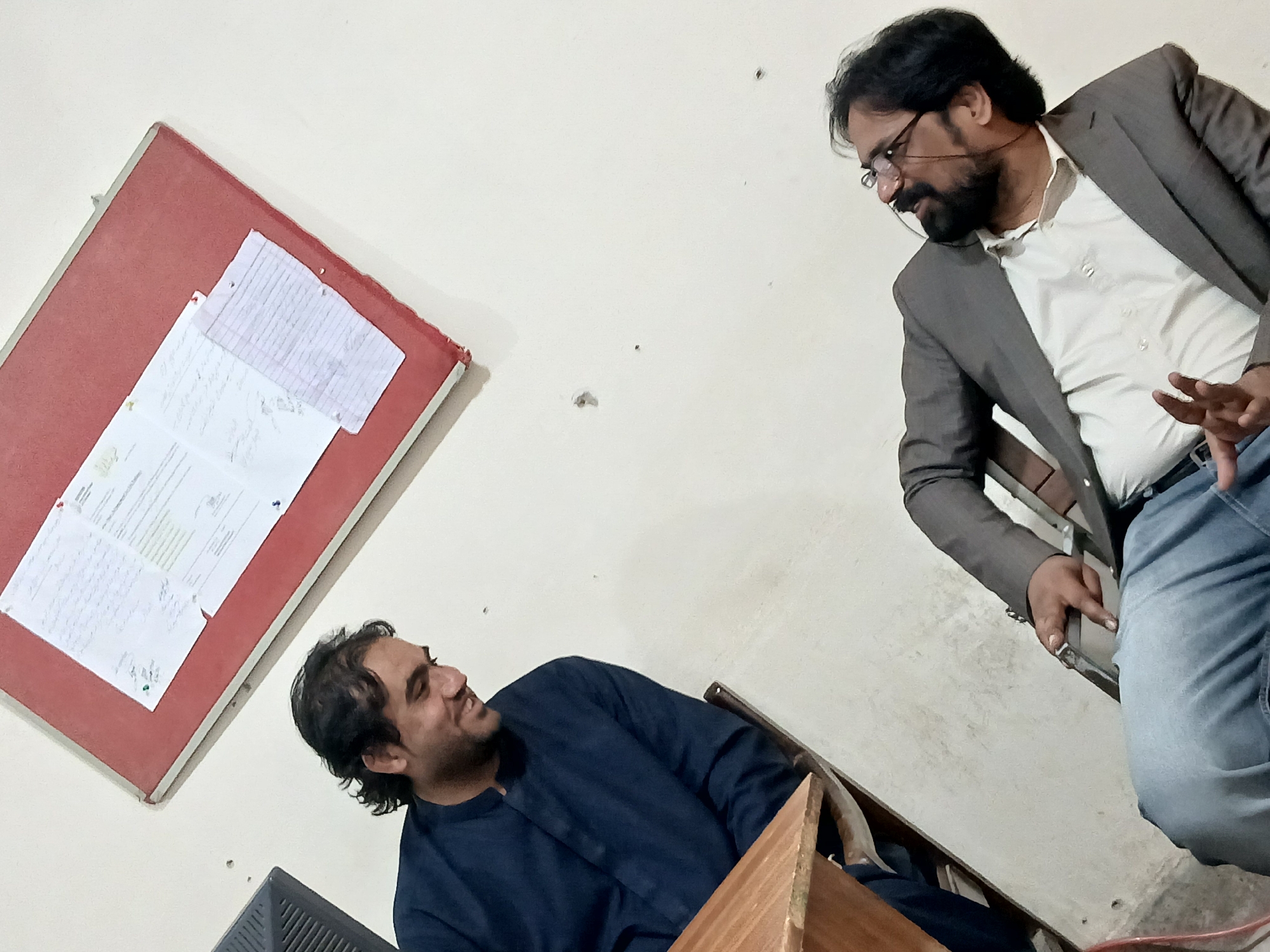 Two men are engaged in a friendly interaction in an indoor setting, likely in Quetta City Tehsil, Pakistan. The scene is illuminated by artificial indoor lighting against a plain white wall.
The man on the right, appearing older, has dark hair, a beard, and wears glasses. He is dressed in a grey suit jacket over a white collared shirt and light blue jeans. He seems to be standing or leaning, gesturing with his left hand, and looking down towards the other man with a slight smile. A thin string from his glasses is visible.
The man on the left, who appears younger, has dark, wavy hair and a beard. He is seated on a wooden chair, wearing a dark blue long-sleeved kurta. He is smiling broadly and looking up at the man on the right. His posture suggests relaxed attention.
To the left of the frame, mounted on the white wall, is a red bulletin board. Several white papers are pinned to it with thumbtacks. These papers contain both handwritten and typed text. Some English words like "document," "instructions," and what appears to be a date format "18-12-2023" are faintly discernible on one document. Other sections show script that resembles Urdu or a similar language, with curved characters.
Other visible objects include parts of a wooden chair and possibly a dark electronic monitor or device towards the bottom left. The overall atmosphere appears casual and positive, suggesting a conversation or discussion between the two individuals.