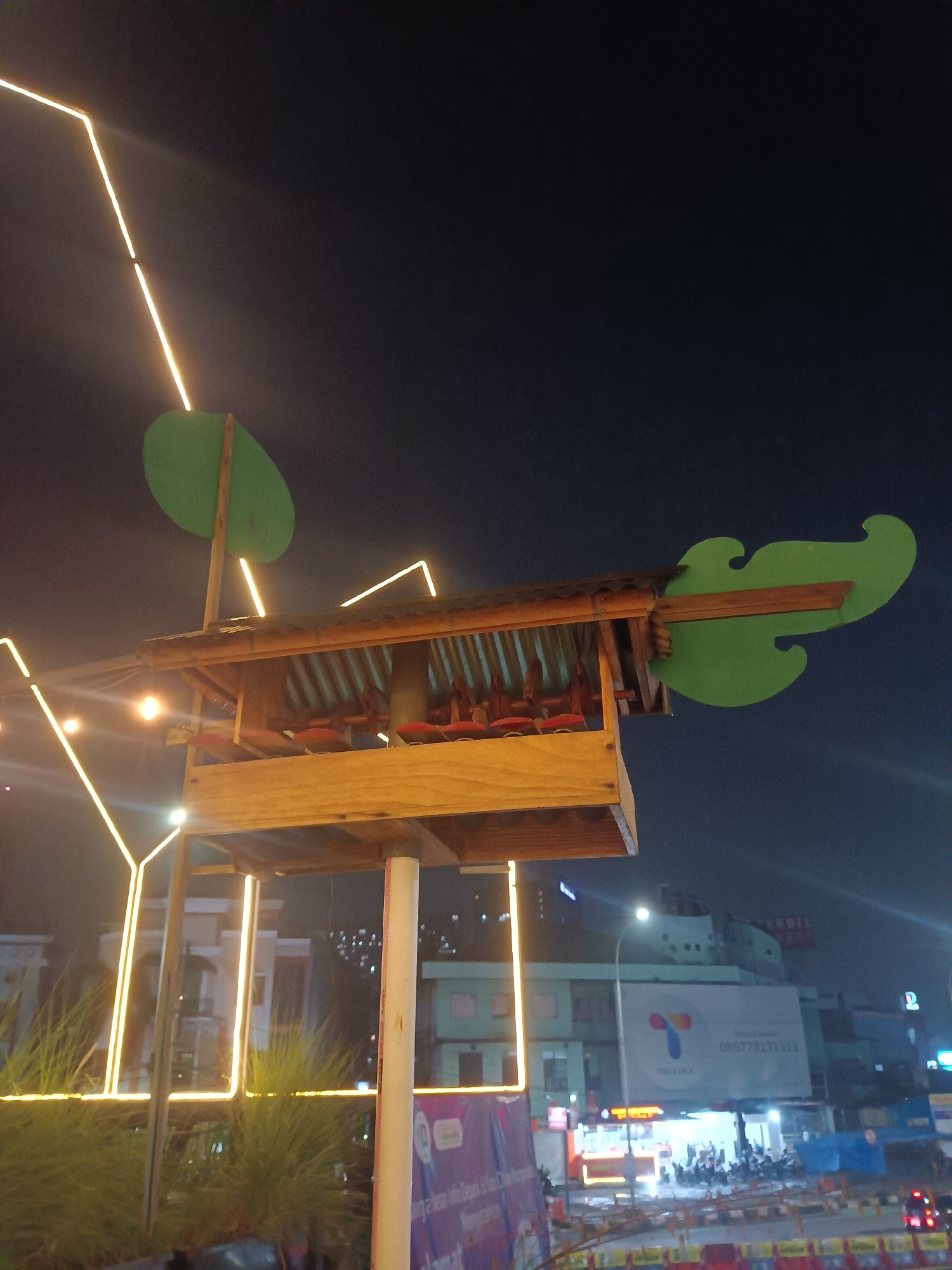 This media file captures a nighttime scene in Depok, Indonesia, featuring a whimsical, open-air structure that resembles a stylized birdhouse or playhouse. The structure, made of wood with a corrugated metal roof, is adorned with large, green, leaf-shaped cutouts, adding a playful, natural element to the urban setting. One leaf extends horizontally to the right, while another stands vertically to the left, resembling a tree. The structure is illuminated by an array of yellow string lights that form geometric patterns in the dark sky, creating a festive and inviting atmosphere.
Below the wooden structure, red seats are visible, suggesting it might be a seating area or a small stage. The foreground is partially obscured by lush, green foliage, likely decorative grass or plants, further enhancing the natural theme. In the background, city lights glow, indicating a bustling urban environment. Buildings and billboards are faintly visible, with one prominent billboard displaying a logo and a phone number, "085773131313". The overall mood is cheerful and inviting, with the ambient light creating a magical, almost dreamlike quality. The time of day is clearly night, and the weather appears clear. The combination of the stylized structure, the colorful lights, and the urban backdrop suggests this might be a public space, perhaps a park, a market, or an outdoor dining area, designed to be visually engaging after dark.