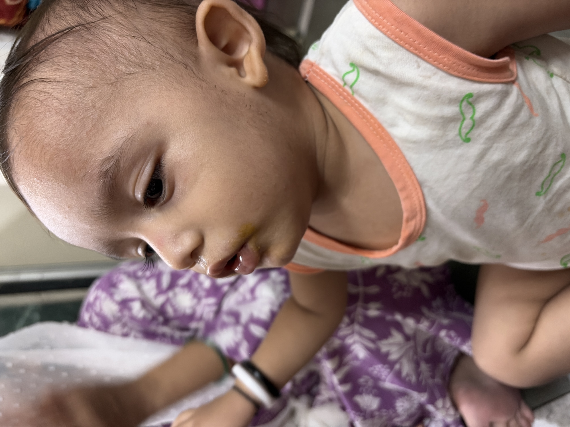 A baby is seated on a floral-patterned purple and white cloth. The baby wears a white tank top with peach trim and a green mustache design. The baby has short dark hair, a fair complexion, and is looking downward. There appears to be some yellow discoloration around the baby's mouth. The baby is wearing a black and white bracelet on its left wrist. The location appears to be indoors. No definitive details place the image in Mumbai, India.