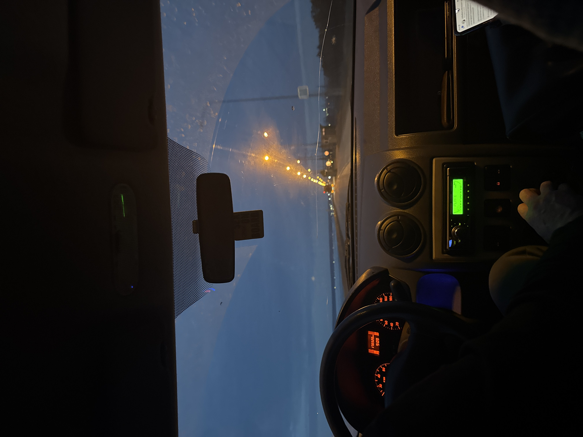 An interior view of a vehicle driving on a road at dusk or night. Outside the windshield, streetlights illuminate the road stretching into the distance. The rearview mirror is visible, along with the car's dashboard, steering wheel, and speedometer, which displays a reading. A person's hand is visible near the car's radio and climate controls. The radio display is illuminated in green. A phone is placed on the dashboard.
The location is potentially Noviy, Russia, based on the prompt. However, there is no explicit location confirmation in the image itself.