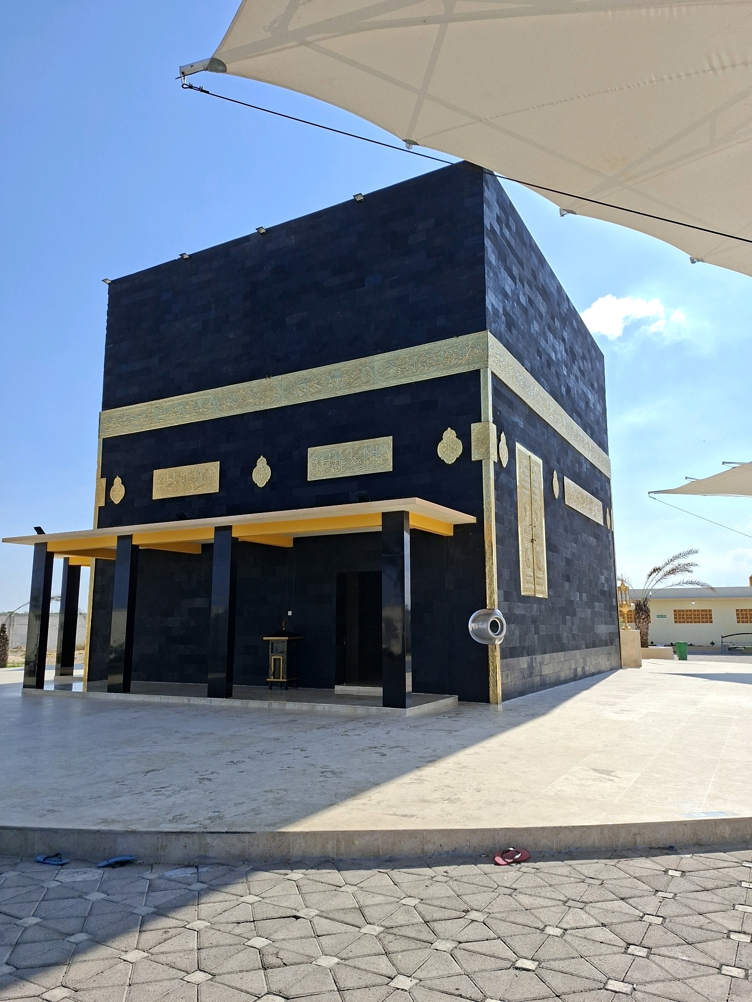 A replica of the Kaaba, a cuboid building, is situated outdoors under a clear blue sky, partially obscured by a large white tensile fabric canopy. The replica is constructed with black brick-like tiles and adorned with golden decorative bands and inscriptions. It features a dark entryway and a covered portico supported by black pillars with a yellow ceiling. The ground surrounding the structure is paved with light-colored square tiles, transitioning to a patterned tile surface in the foreground where a pair of blue flip-flops and a single red flip-flop are visible. In the background, a low building with green and white accents and a palm tree can be observed, along with some other ambient structures and vegetation. The scene appears to be in Jrengik, Indonesia.