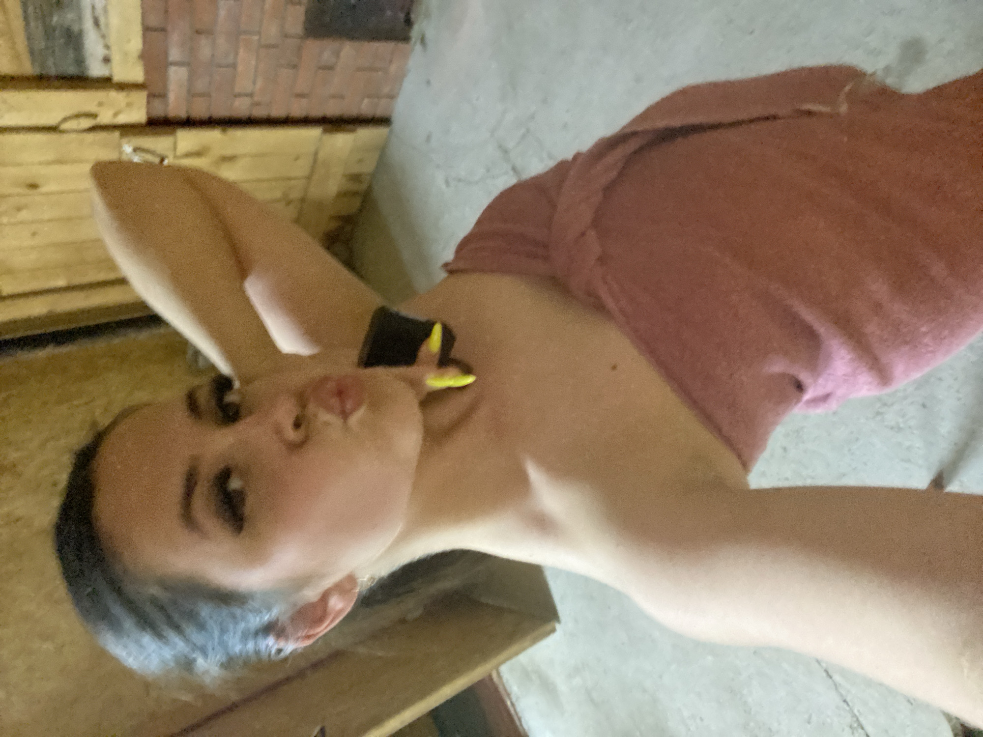 A woman is posing for a selfie. She has dark hair and makeup. She is wearing a pink garment and a black watch with a yellow strap. The background includes wooden surfaces and a concrete-like floor. There is no evidence indicating the location is Zhigalki, Russia.