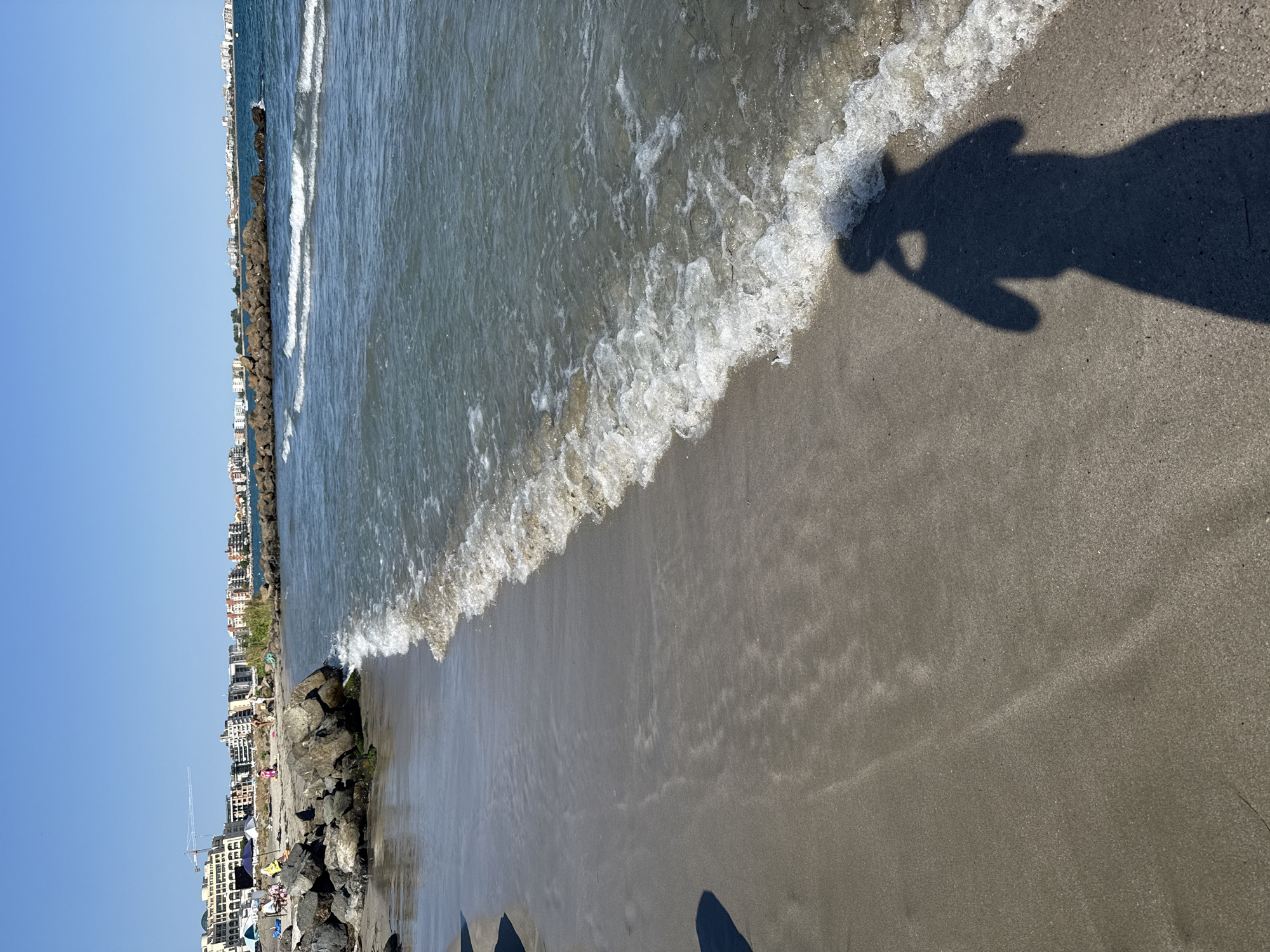 The image captures a serene beach scene in Pomorie, Bulgaria, on a clear, sunny day. The foreground is dominated by the wet sand of the shoreline, where gentle waves are lapping at the beach, leaving behind a foamy white crest. The water itself is a translucent blue-green, with sunlight glinting off its surface. In the mid-ground, a rocky breakwater juts out into the sea, its dark stones contrasting with the lighter sand and water. Beyond the breakwater, the coastline of Pomorie stretches into the distance, characterized by a series of modern, multi-story buildings, likely hotels or apartment complexes, painted in various light colors, predominantly white and beige. A few scattered umbrellas and beach chairs are visible on the sandy area further up the beach, suggesting a few people are enjoying the seaside. A prominent dark shadow of a person holding a camera is cast on the sand in the lower right corner of the image, indicating the photographer's presence. The sky above is a cloudless, bright blue, completing the idyllic coastal panorama.