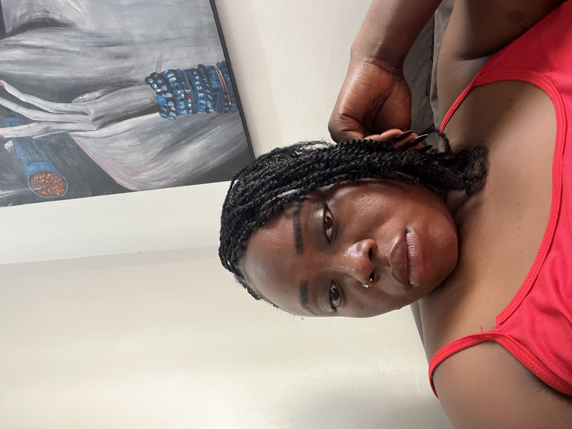 A close-up, waist-up, eye-level, indoor photograph shows a Black woman with braided hair and a red top. She is lying down, with her head resting on her right arm and hand. Her left arm is bent at the elbow and her hand is resting near her hair. She is looking directly at the camera with a neutral expression. Behind her, on the wall, is a painting of a torso with a hand adorned with bracelets. The background is a plain white wall, and she appears to be on a couch or bed. No information is available to determine the specific location in Abuja, Nigeria.