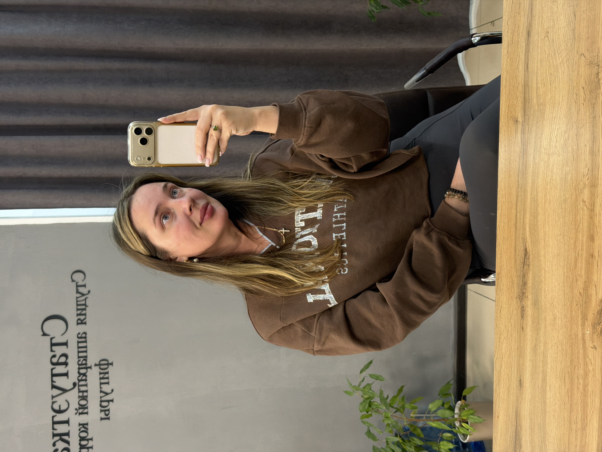 A woman is captured in a selfie taken in a mirror or reflective surface, indicated by the reversed text on the wall. She is positioned on the right side of the frame, seated in a dark chair, and appears to be looking towards the camera with a subtle upward gaze and a slight smile. She has long, light brown hair with blonde highlights, blue eyes, and fair skin.
She is wearing a brown crewneck sweatshirt with white, reversed text on the front, which includes the clear letters "T" and "THE". Around her neck is a delicate gold chain with a cross pendant. On her left hand, which holds the phone, she wears a ring with a prominent green stone. Her right wrist features beaded bracelets. Her lower body is clad in dark gray or black leggings.
She holds a gold or beige iPhone with a clear protective case, showcasing its triple camera setup, in her left hand, extended towards the reflective surface.
The background features a light gray wall on the left with prominent reversed text. This text reads "Студия Эпиляции и косметологии" (Studio of Epilation and Cosmetology) and "ФилАВРИ" (FilAVRI), suggesting the location is a beauty or cosmetology studio. Above this wall, dark gray horizontal blinds or curtains are visible. To the right of the woman, a light wooden table or counter surface extends vertically. A small green potted plant is partially visible in the bottom center foreground, adding a touch of nature to the indoor setting. The overall lighting suggests it is daytime. The location is Chaikovsky, Russia.