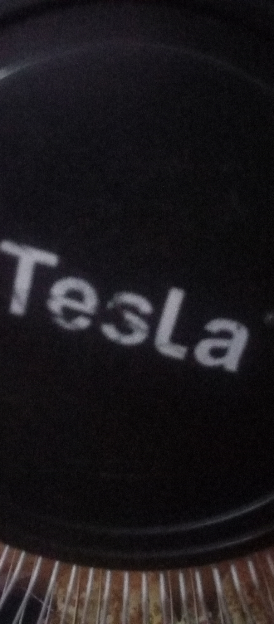The image is a close-up, slightly blurry photograph of the word "Tesla" printed in white, sans-serif font on a dark, possibly black, surface. The surface appears to be textured or possibly fabric-like. The lighting in the image is uneven, with a bright glare on the upper left portion of the surface, which casts subtle shadows on the lettering. The bottom of the image shows a ribbed, metallic or plastic surface, suggesting the object might be a part of a larger item, possibly an air filter or a speaker grille. There are no people or other identifiable objects in the scene. The image does not provide any information or context related to the city of Lubuk Pakam, Indonesia, or any urban environment.