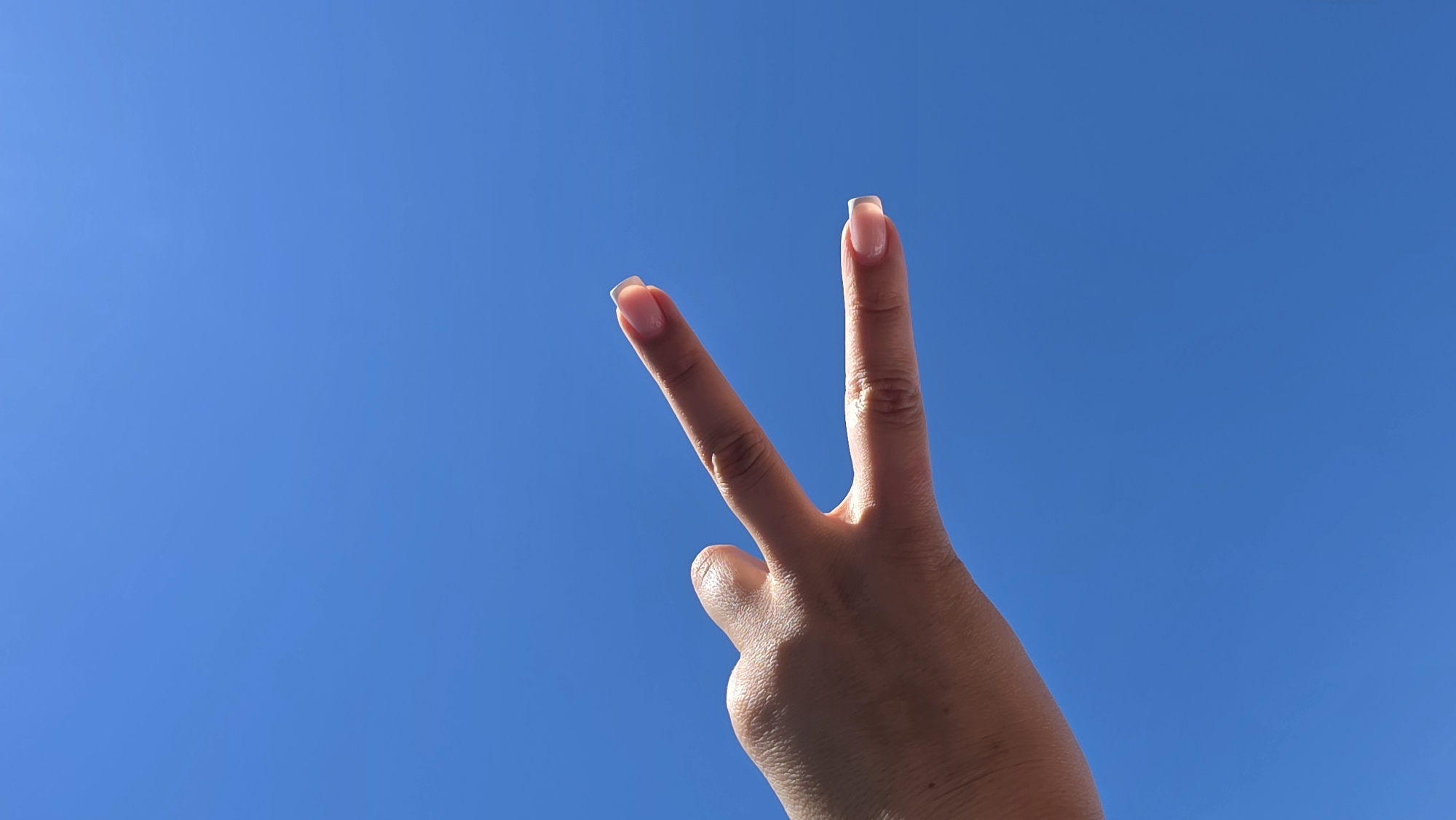 A close-up, low-angle outdoor shot features a person's hand making a peace sign against a clear, bright blue sky. The hand, with manicured nails, extends from the left side of the frame, with the index and middle fingers held up in a V-shape, while the thumb and other fingers are tucked in. The lighting suggests it is daytime. The image was captured in Pivdenne, Ukraine.